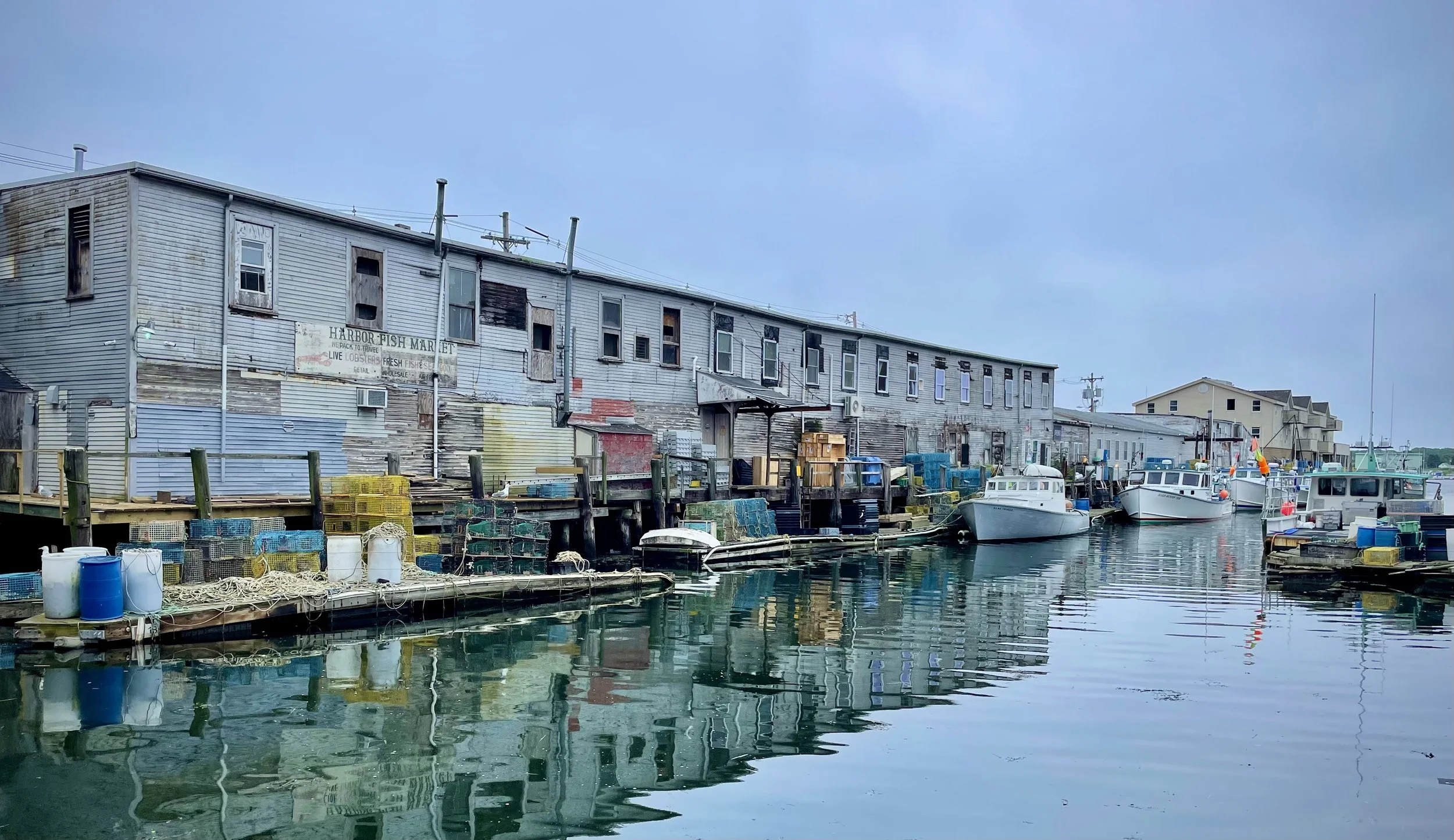 A waterfront scene featuring an old, weathered building with a sign for Harbor Fish Market, boats docked along the pier, crates and barrels on the dock, and water reflecting the scene under a cloudy sky.
