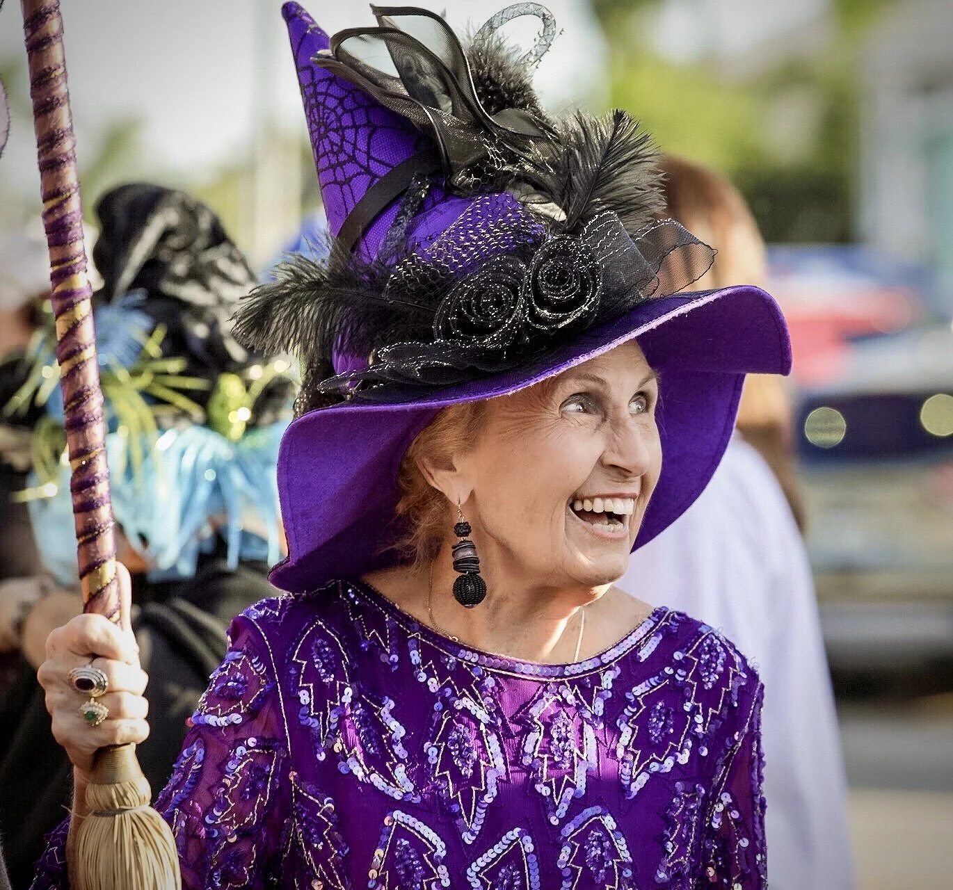 An elderly woman wearing a purple hat decorated with black feathers, black spiderweb accents, and a veil, smiling and holding a stick with a braid, dressed in a purple sequined top with intricate patterns, at an outdoor event.