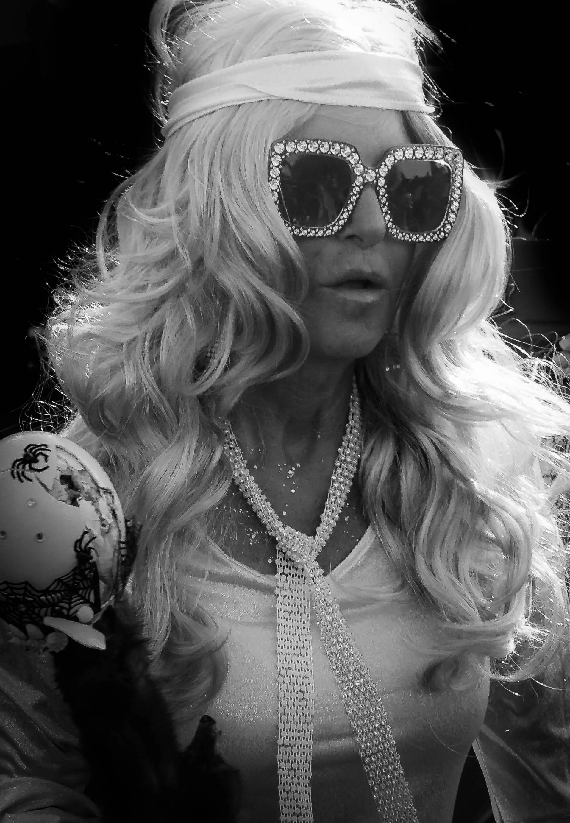 A woman with long wavy blonde hair wearing large, rhinestone-encrusted sunglasses, a white headband, and a shiny top. She is accessorized with layered necklaces and is holding a fan with a floral pattern. The photo appears to be taken outdoors.