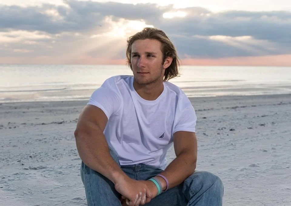 A young man with long blond hair sitting on a sandy beach during sunset, wearing a white t-shirt and blue jeans, looking to the side.