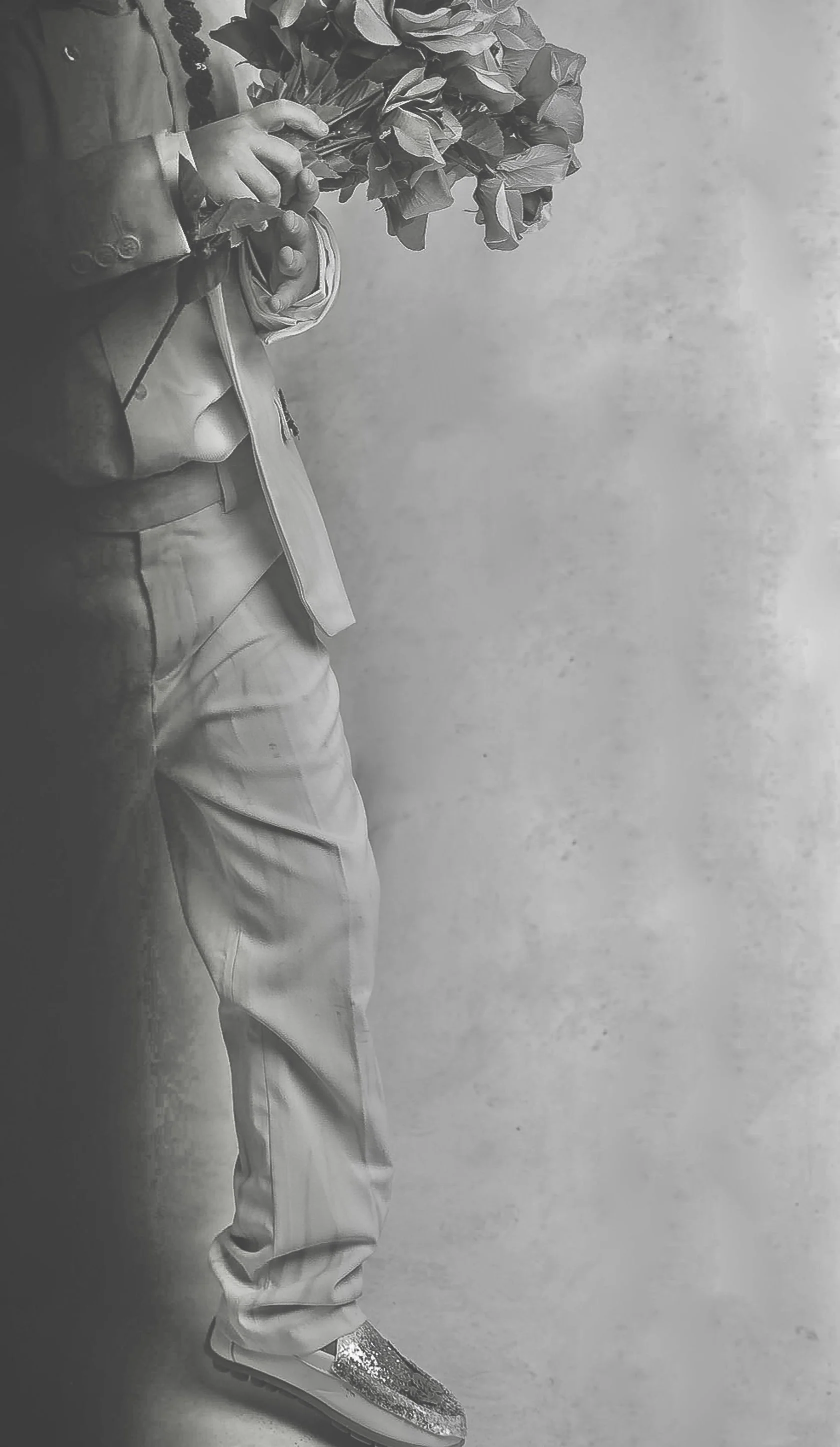 Person dressed in light-colored pants, jacket, and sneakers holding a large bouquet of flowers against a plain background.