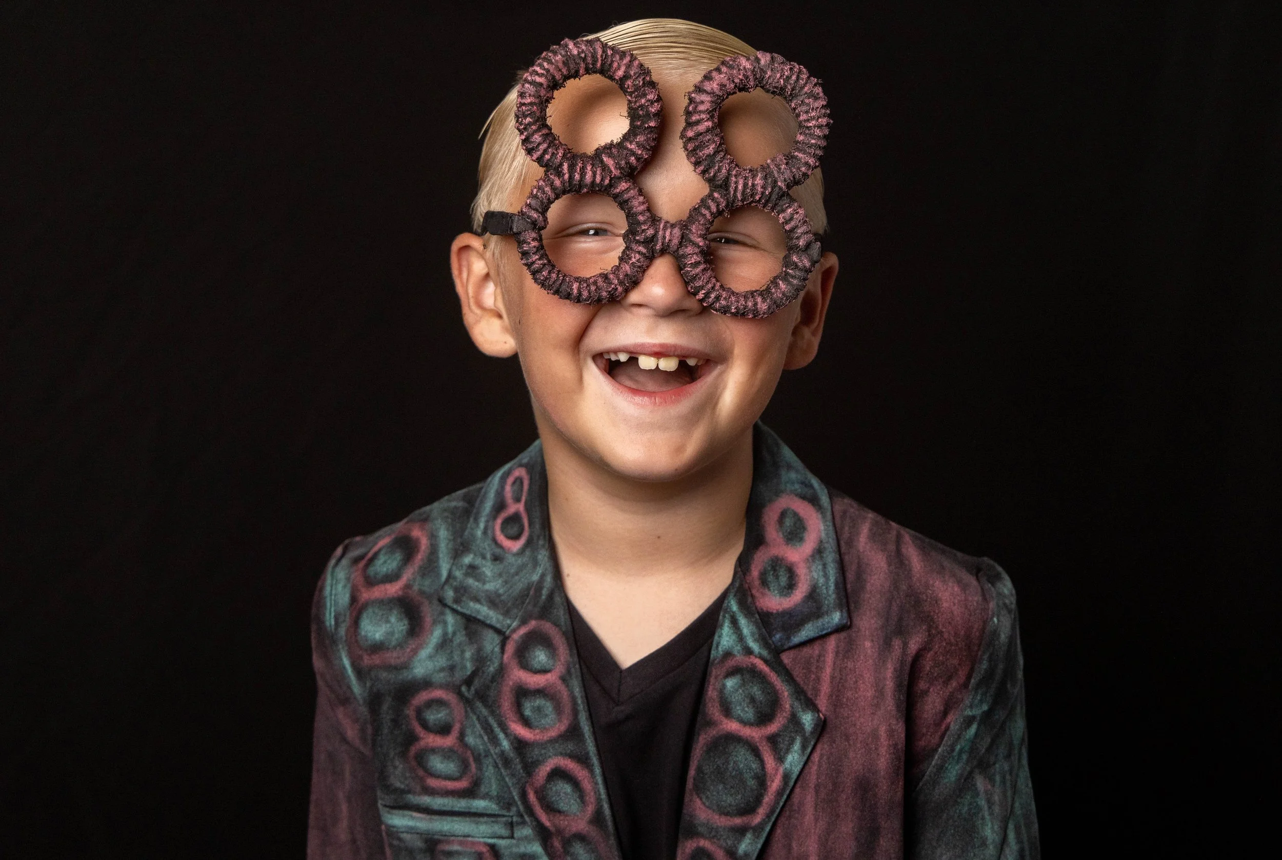 Young boy with missing front teeth, wearing large circular glasses and a patterned jacket, smiling against a black background.