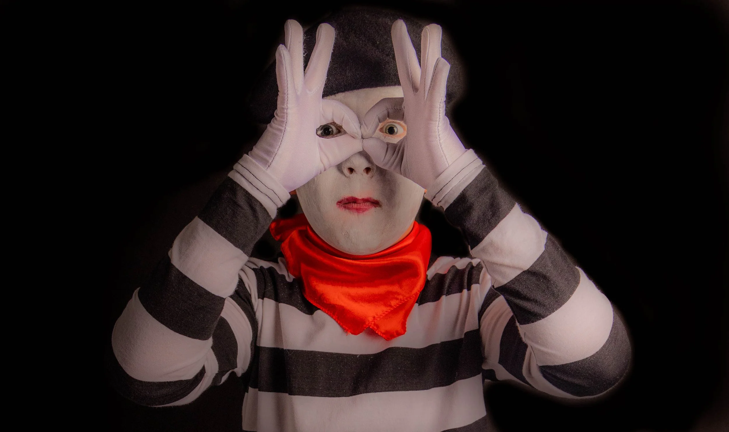 A person dressed as a mime is making a camera frame with their hands over their face. The mime is wearing white gloves, a black and white striped shirt, a red bandana, and a black hat. They are set against a dark background.