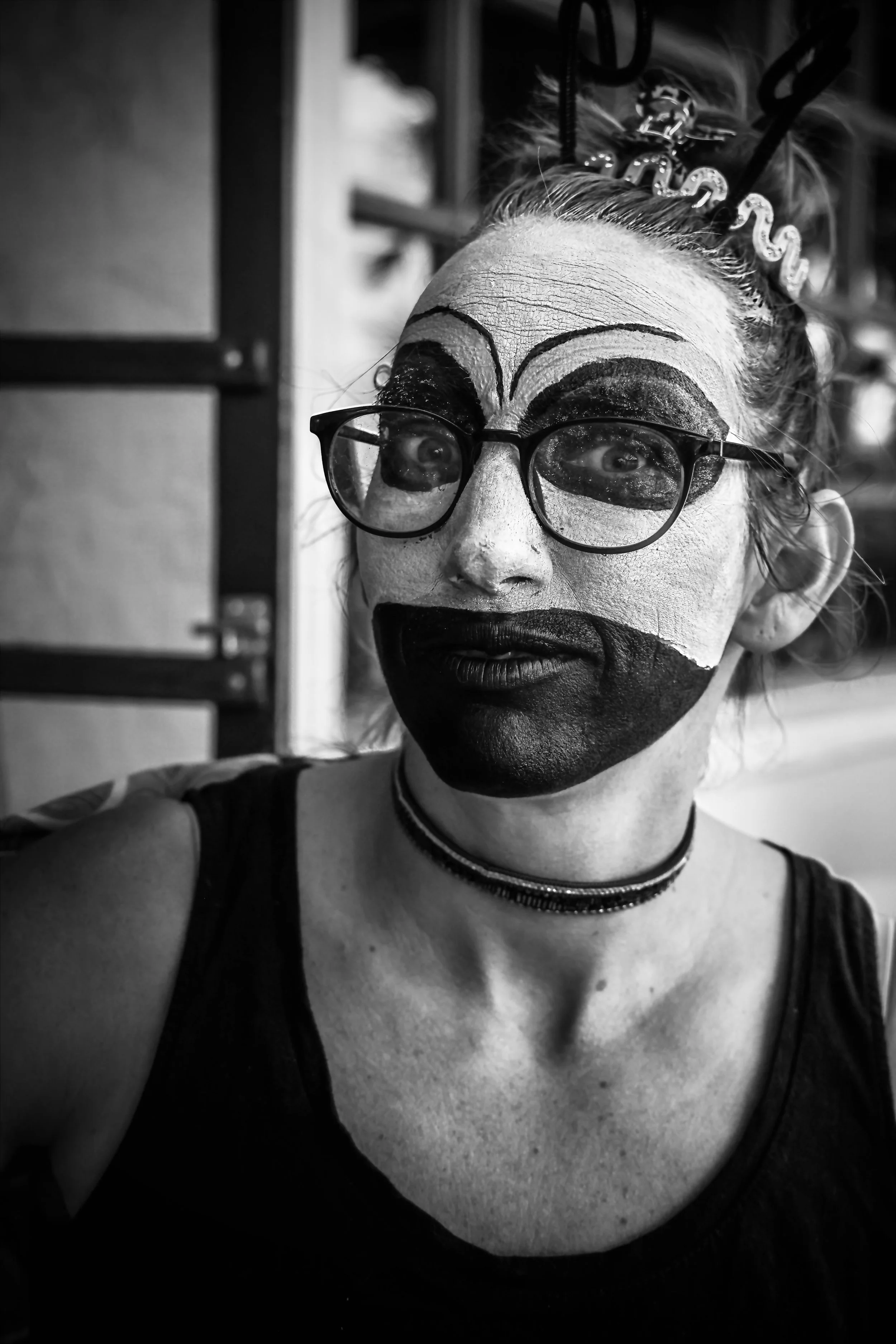 A person with clown makeup and glasses, wearing a necklace, taking a selfie in black and white. Their face is painted white with large black and white designs around the eyes and lips, and a headband with the word 'MISS' on it.