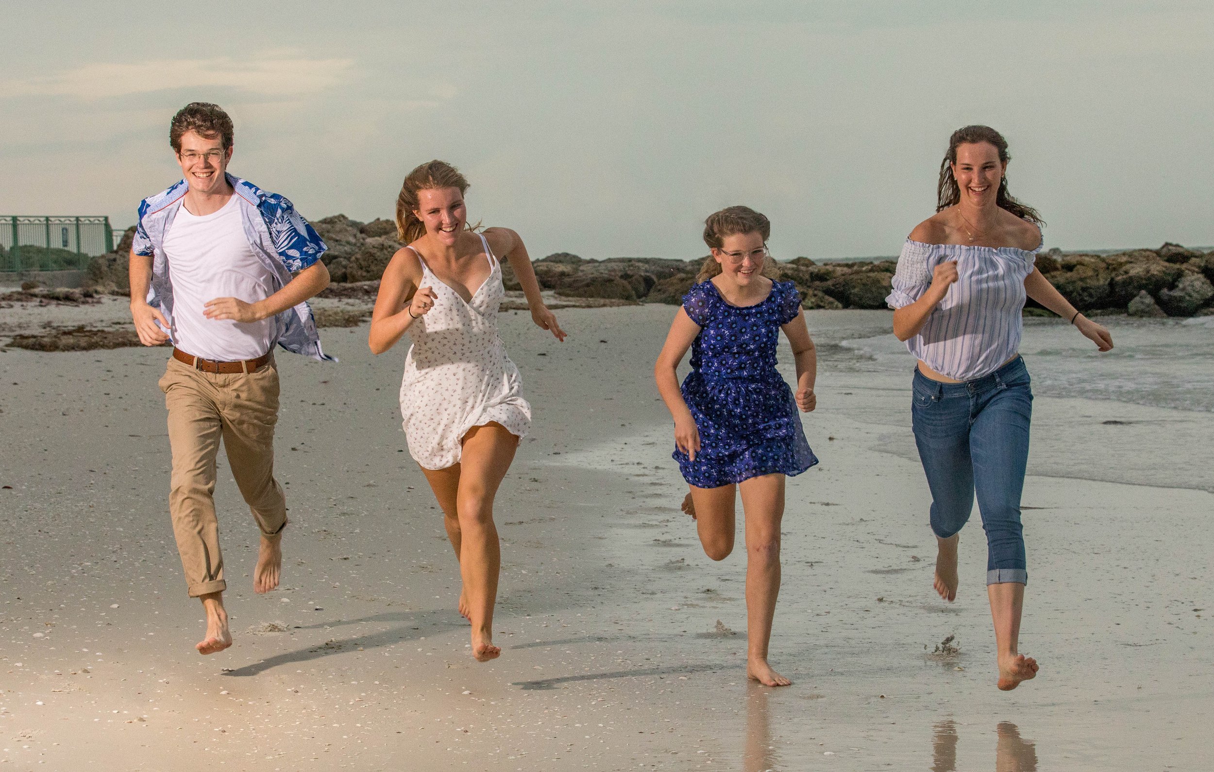 Group of five young women and men running barefoot on the beach along the shoreline, smiling and enjoying themselves.