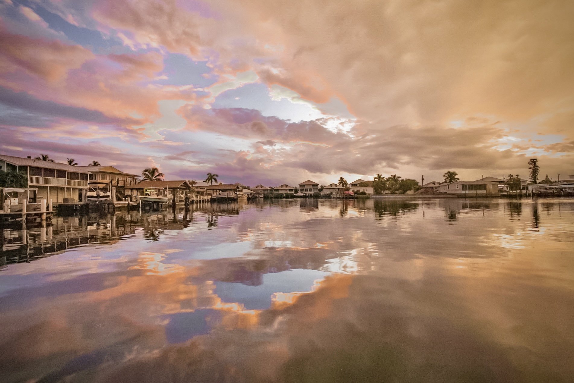 A calm waterway reflecting a colorful sky at sunset, with houses and boats along the shoreline and palm trees in the background.
