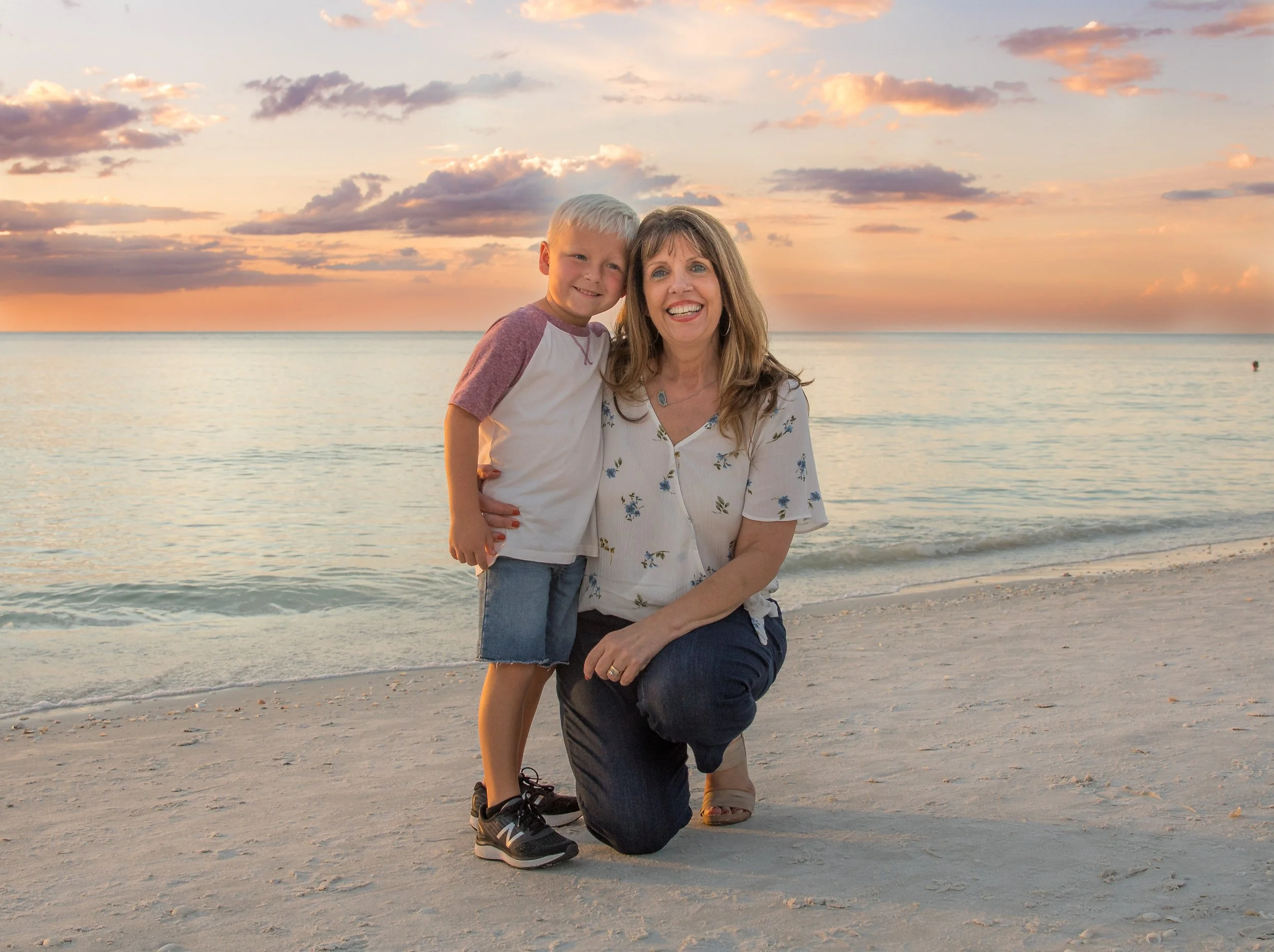 A smiling woman and a young boy at the beach during sunset. The woman is kneeling on one knee, and the boy is standing close to her with his arm around her shoulders. The sky is partly cloudy with pink and orange hues, and the calm water stretches ou