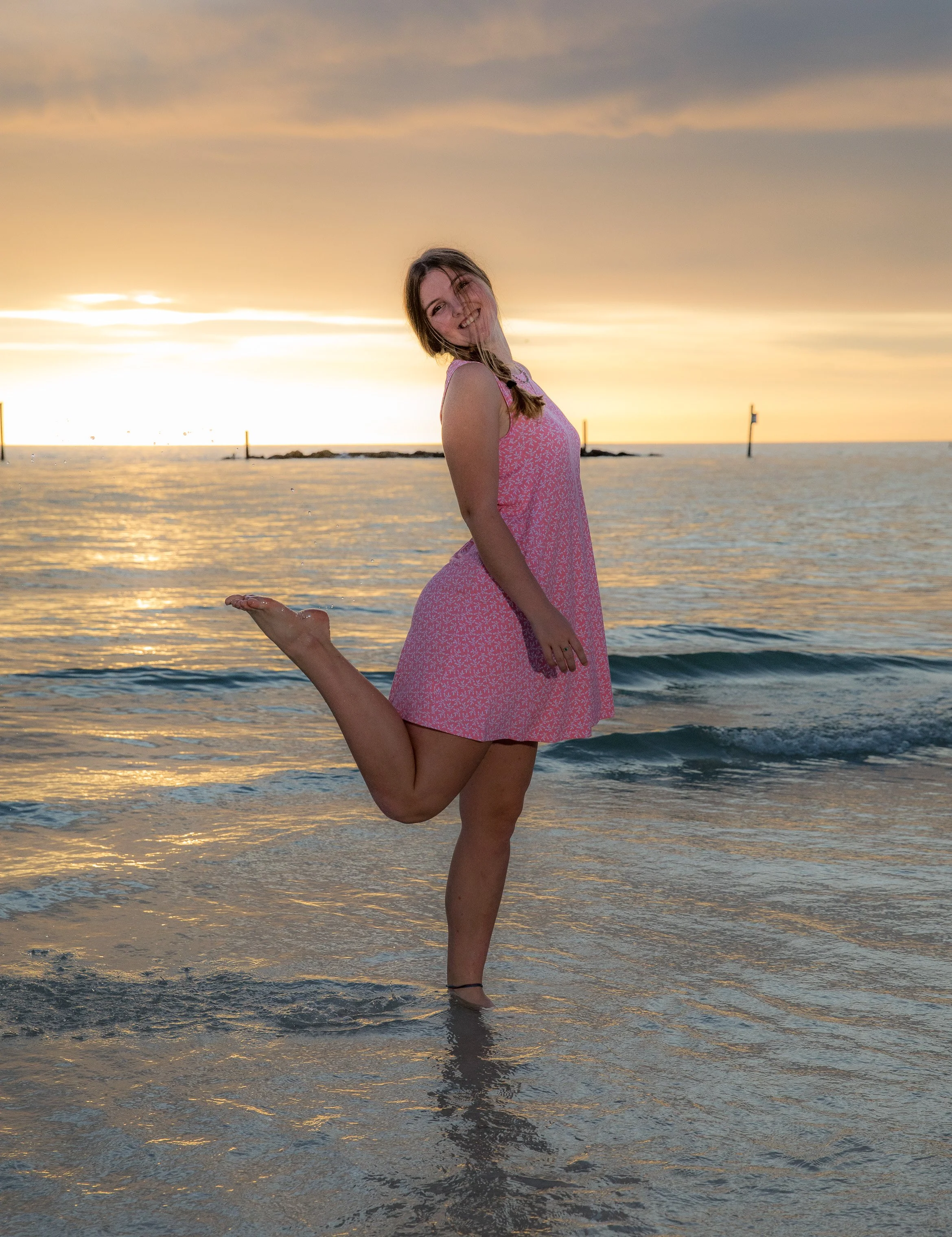 A woman in a pink dress is standing on one leg in shallow water at the beach during sunset, balancing on her other leg with her foot held by her hand.