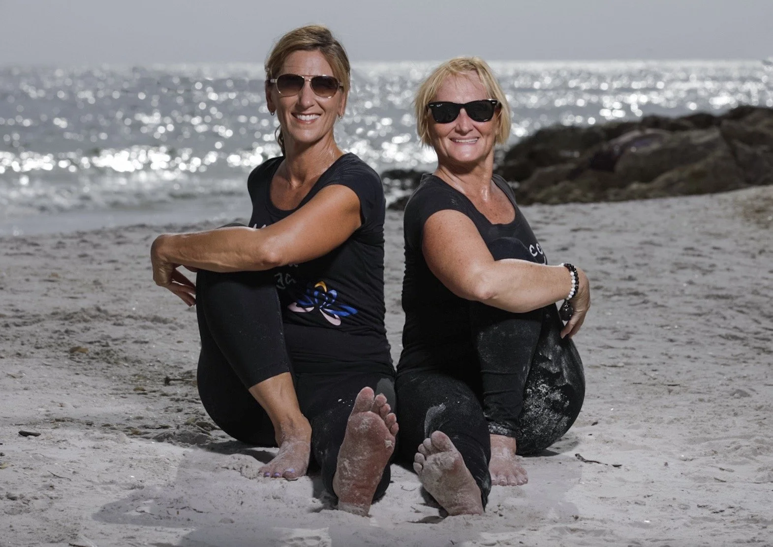 Two women sitting on a sandy beach, back to back, smiling at the camera, with the ocean and rocks in the background, both wearing black clothes and sunglasses.