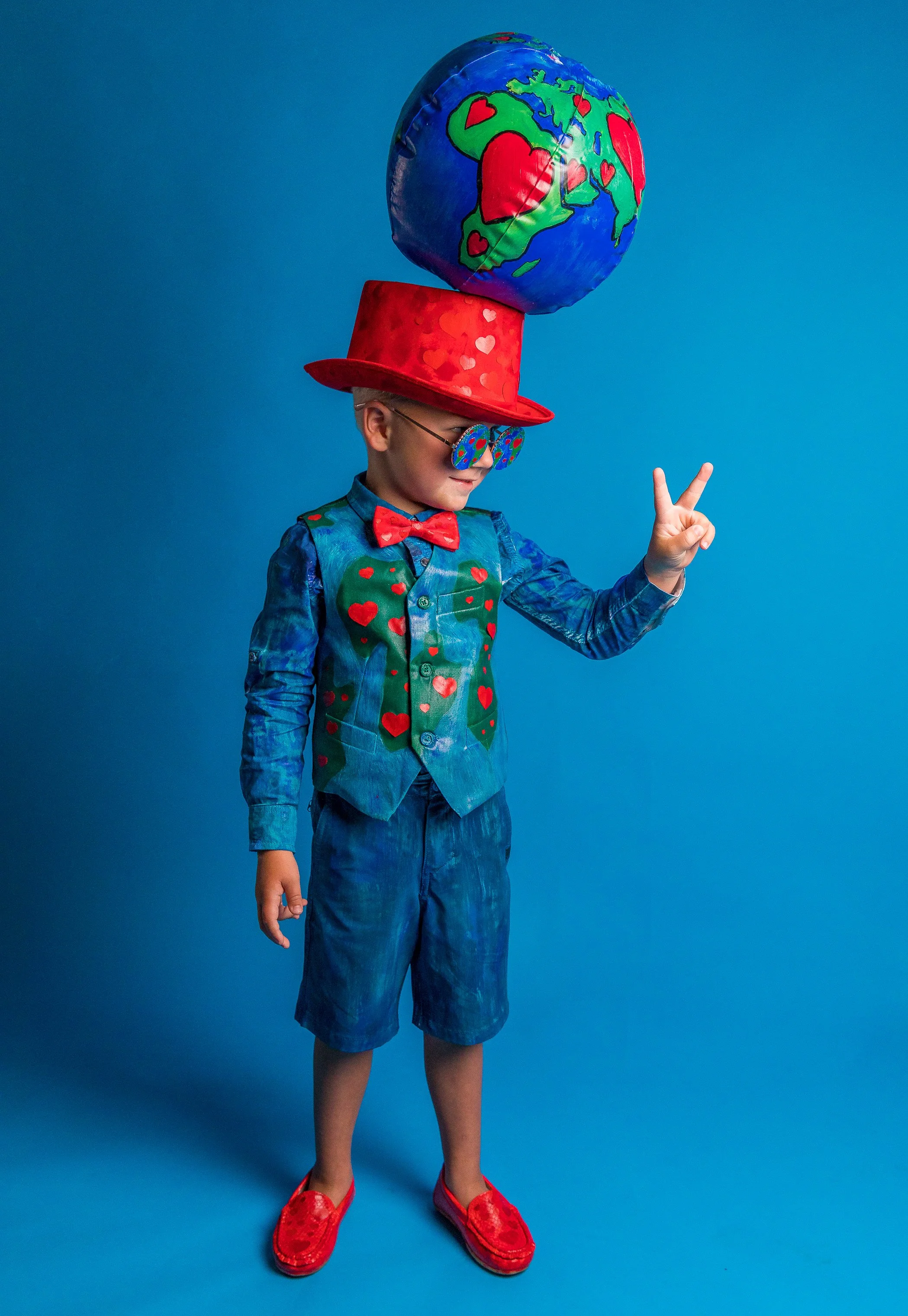 Child dressed in colorful clothing and accessories standing against a blue background. The child is wearing a red hat with hearts, sunglasses, a blue shirt with a matching vest and bow tie, denim shorts, and red shoes. They are balancing a balloon sh