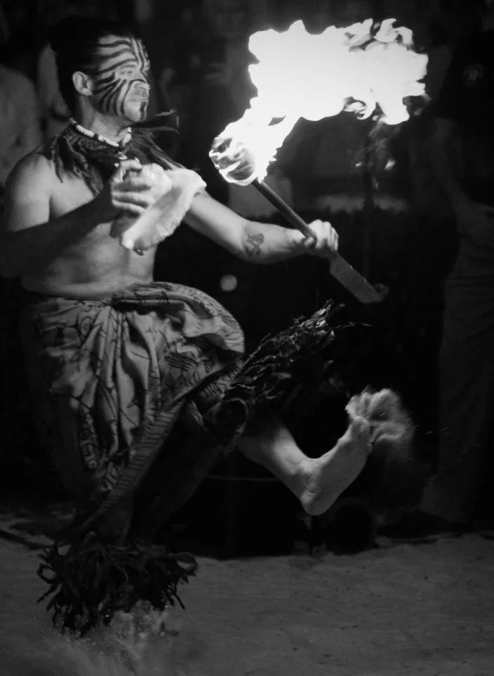 Indigenous man performing fire dancing with a flaming torch, face painted in traditional patterns, wearing patterned cloth, barefoot, with a feathered object in one hand, in a dark setting.