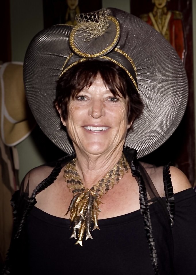 A woman smiling, wearing an elaborate large hat with gold accents and a netted decoration, and a statement necklace with gold leaf details.