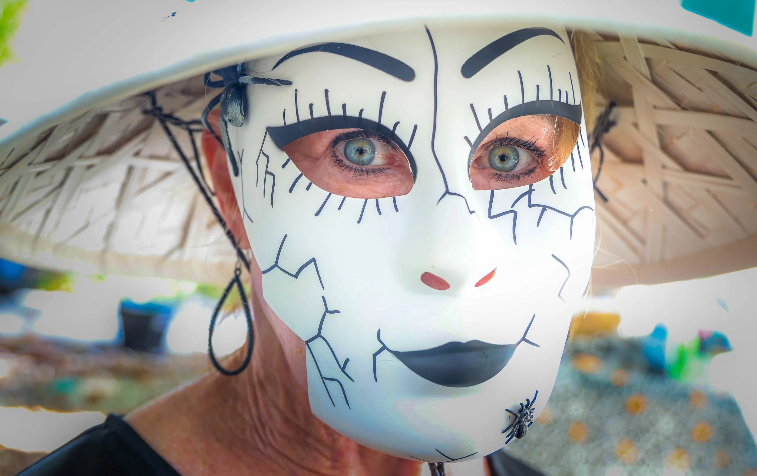 Person wearing a white mask with black lines and eye holes, peeking out from underneath a conical hat, outdoors during daytime.