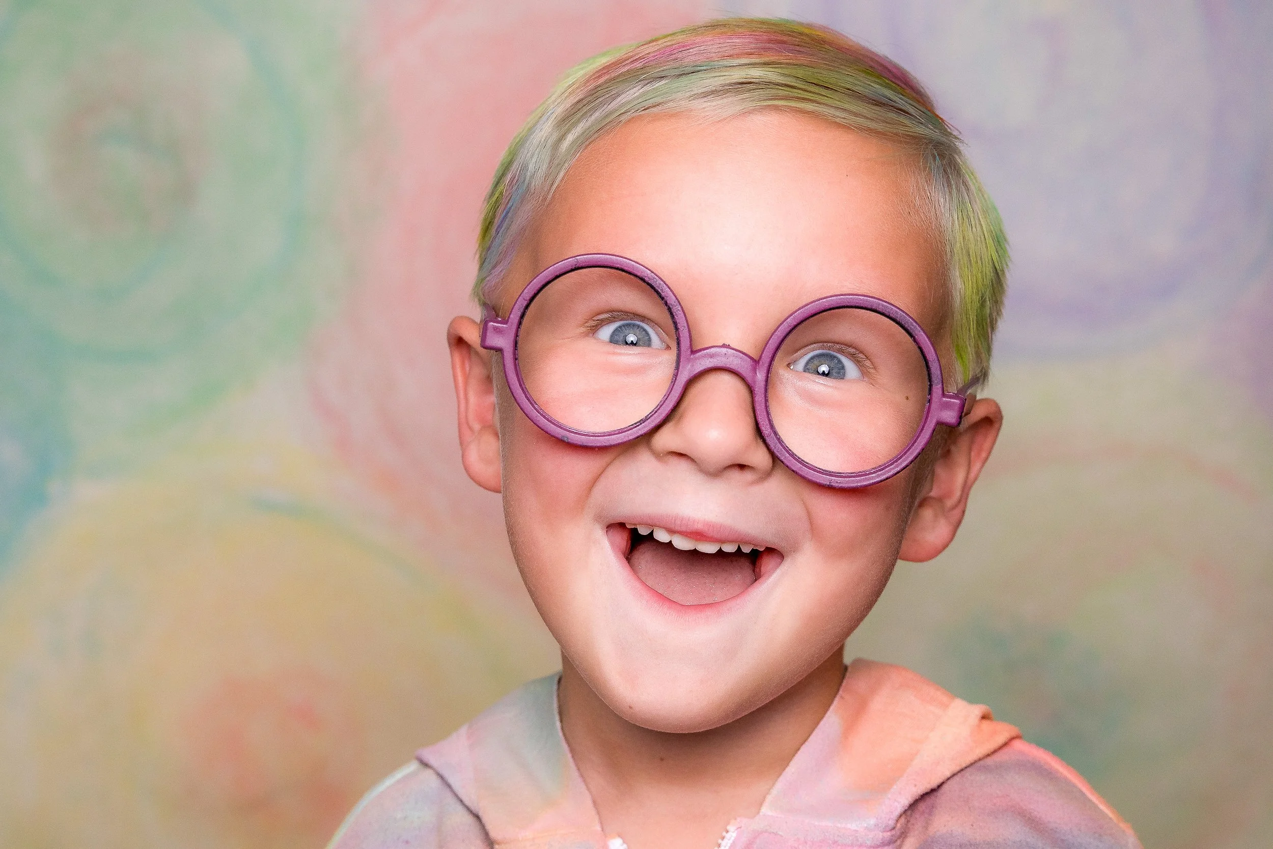 A young boy with short blond and pastel-colored hair, wearing large purple round glasses, smiling widely with his mouth open, against a pastel, watercolor-like background.