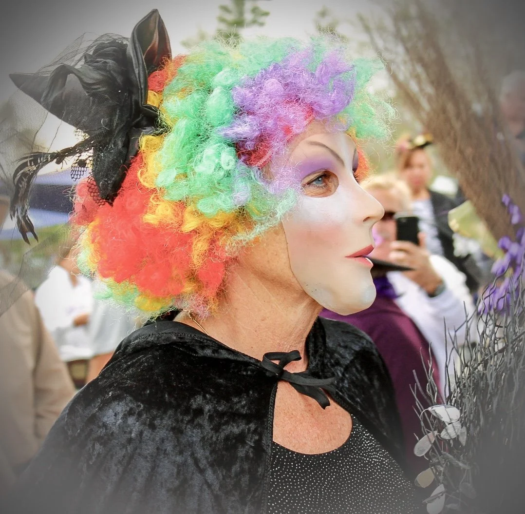 Person dressed as a clown with rainbow-colored curly wig, white face makeup, colorful eyeshadow, and a black cape, holding branches with purple flowers at an outdoor event.