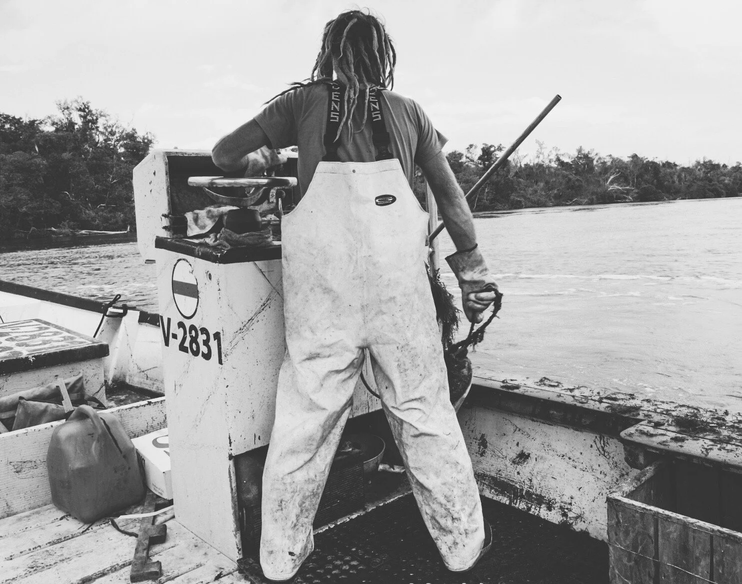 A person with dreadlocks in a raincoat on a boat, holding a fishing net, with water and trees in the background.