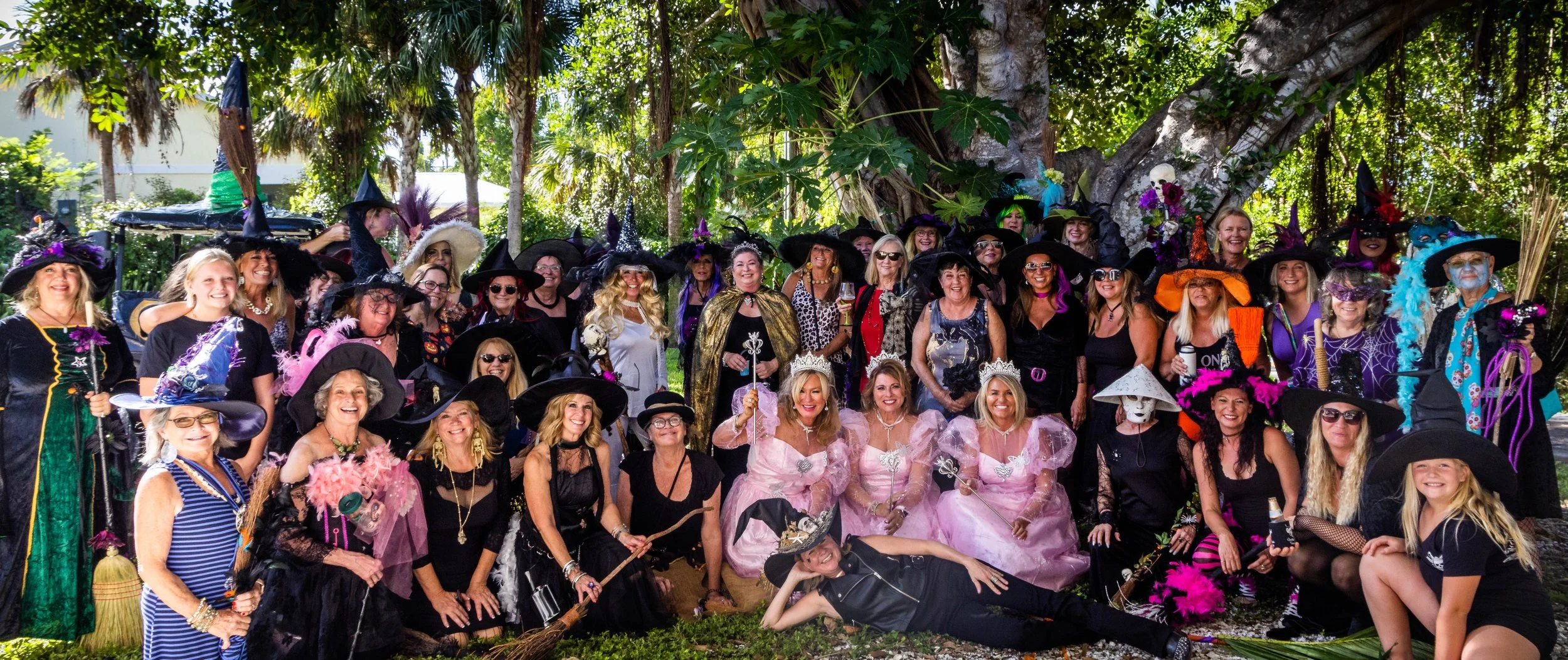Large group of women and a few children dressed as witches, princesses, and fairies, wearing colorful costumes and witch hats, gathered outdoors in front of a large tree with lush green foliage, celebrating a themed event.