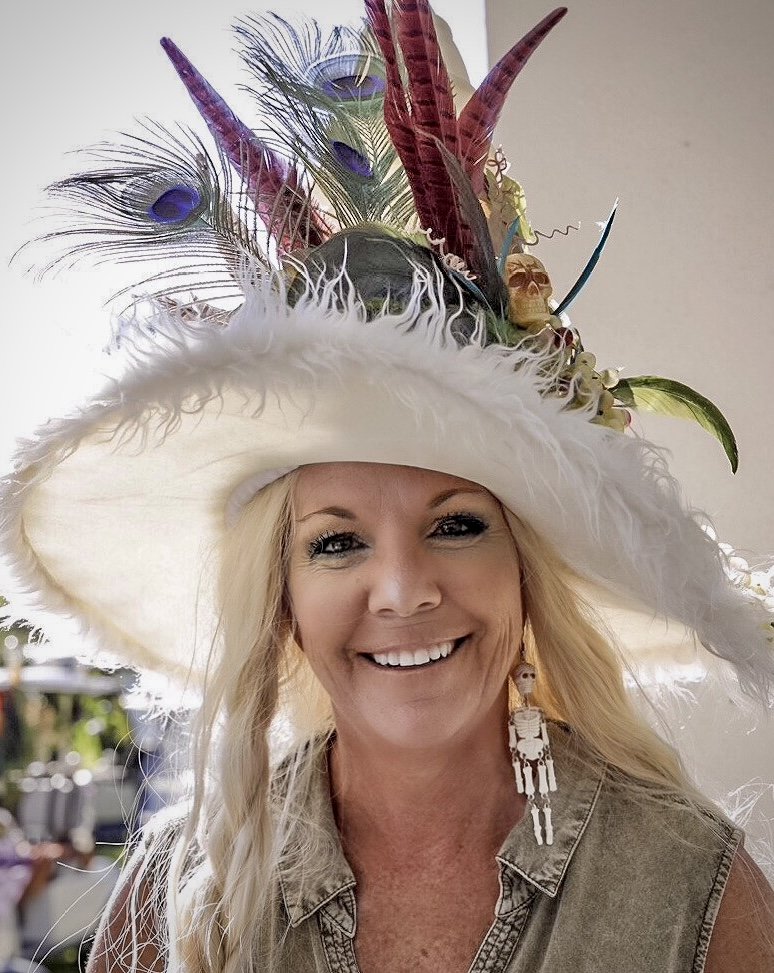 Woman smiling while wearing an elaborate white feathered hat decorated with peacock feathers, colorful feathers, and various embellishments.