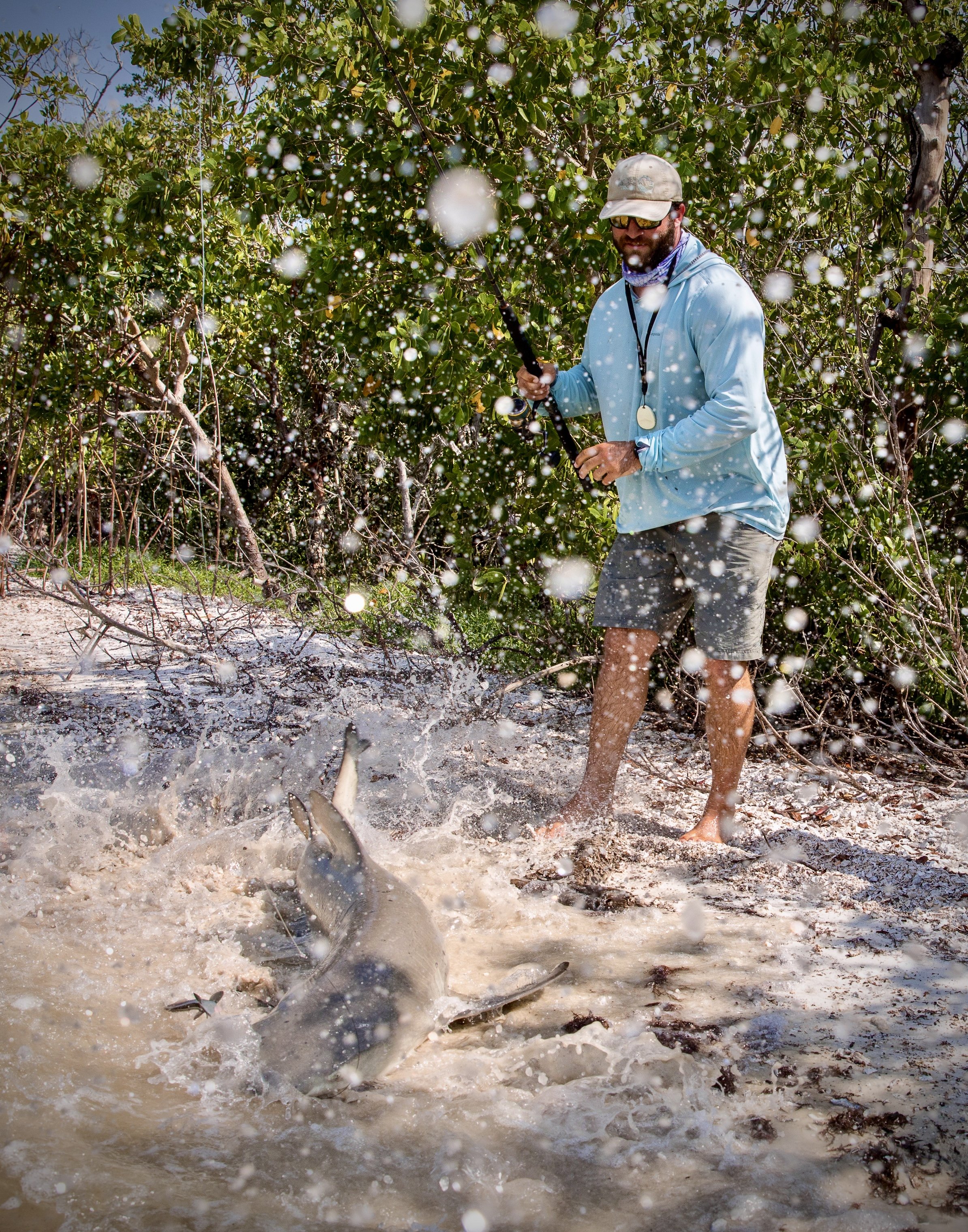 A man wearing a hat, sunglasses, and a light blue jacket is fishing in a body of muddy water surrounded by dense green bushes. He is holding a fishing pole, and fish are jumping out of the water, creating splashes.