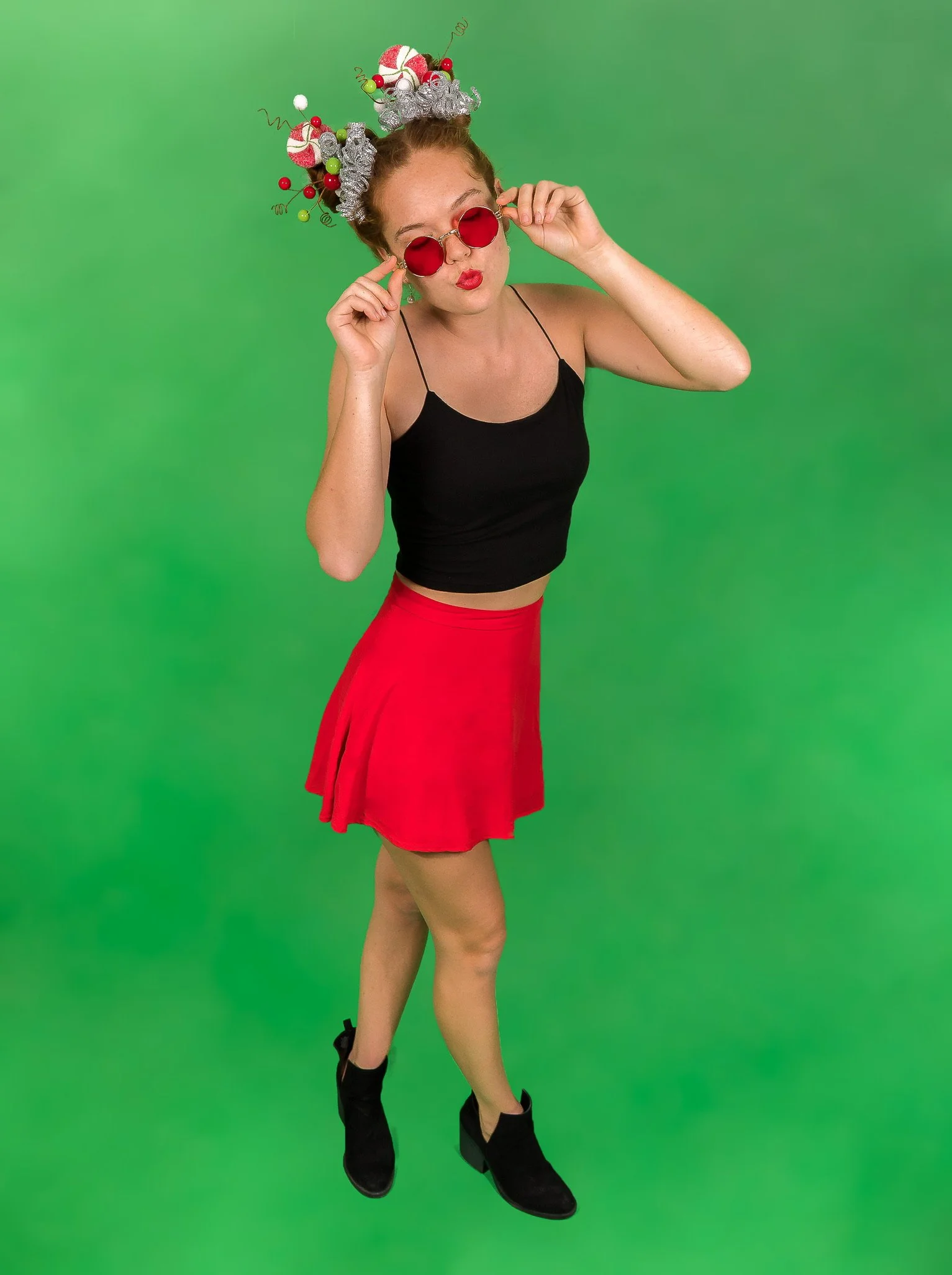 A woman with red hair in a bun wearing red sunglasses, a black spaghetti strap crop top, a red skirt, and black ankle boots stands against a green background. She is adjusting her sunglasses and has a festive headpiece made of holiday-themed decorati