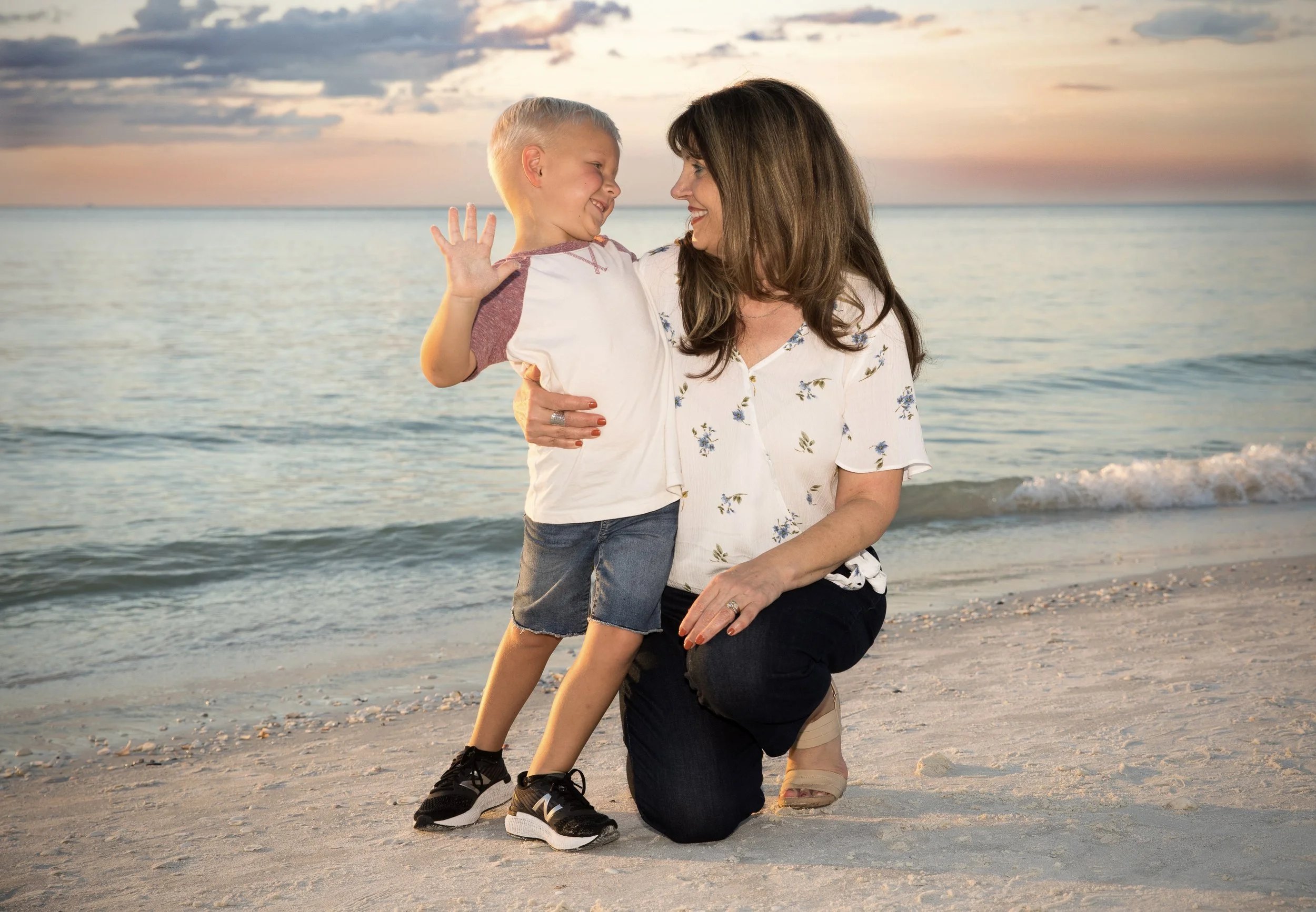A woman kneeling on the beach, holding a smiling young boy on her shoulders during sunset with ocean waves in the background.