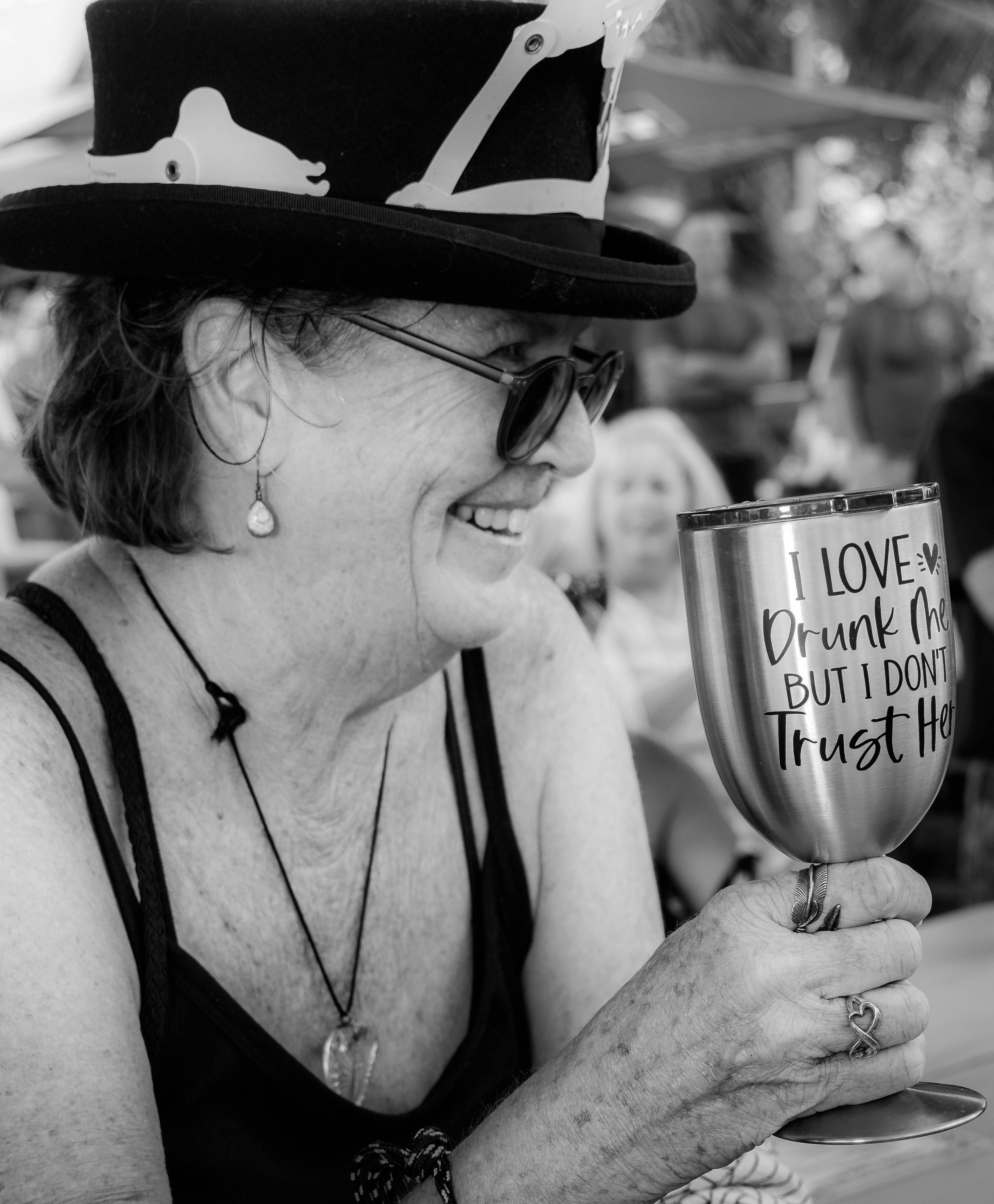 Close-up of a smiling woman with short hair, wearing sunglasses and a black top hat with a rhinoceros design, holding a glass with a drink that has a message, wearing rings and a necklace, with background of trees and outdoor setting.