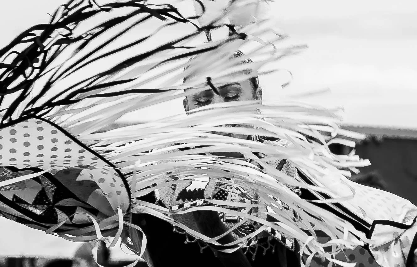 A person partially obscured behind a large, decorative arrangement of ribbons, with only their eyes and forehead visible, in a black and white setting.