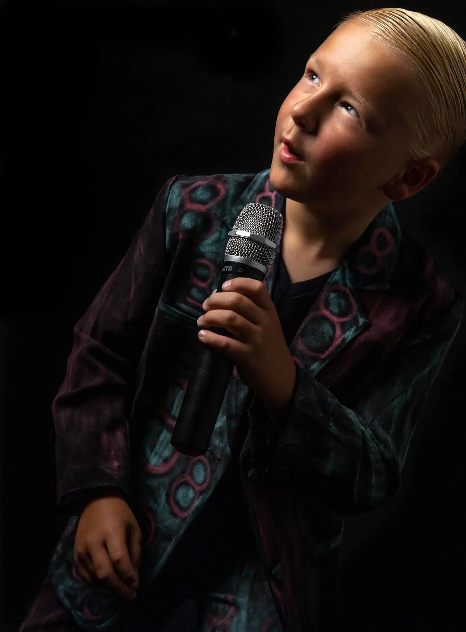 A young person with blonde hair holding a microphone, wearing a colorful jacket, against a dark background.