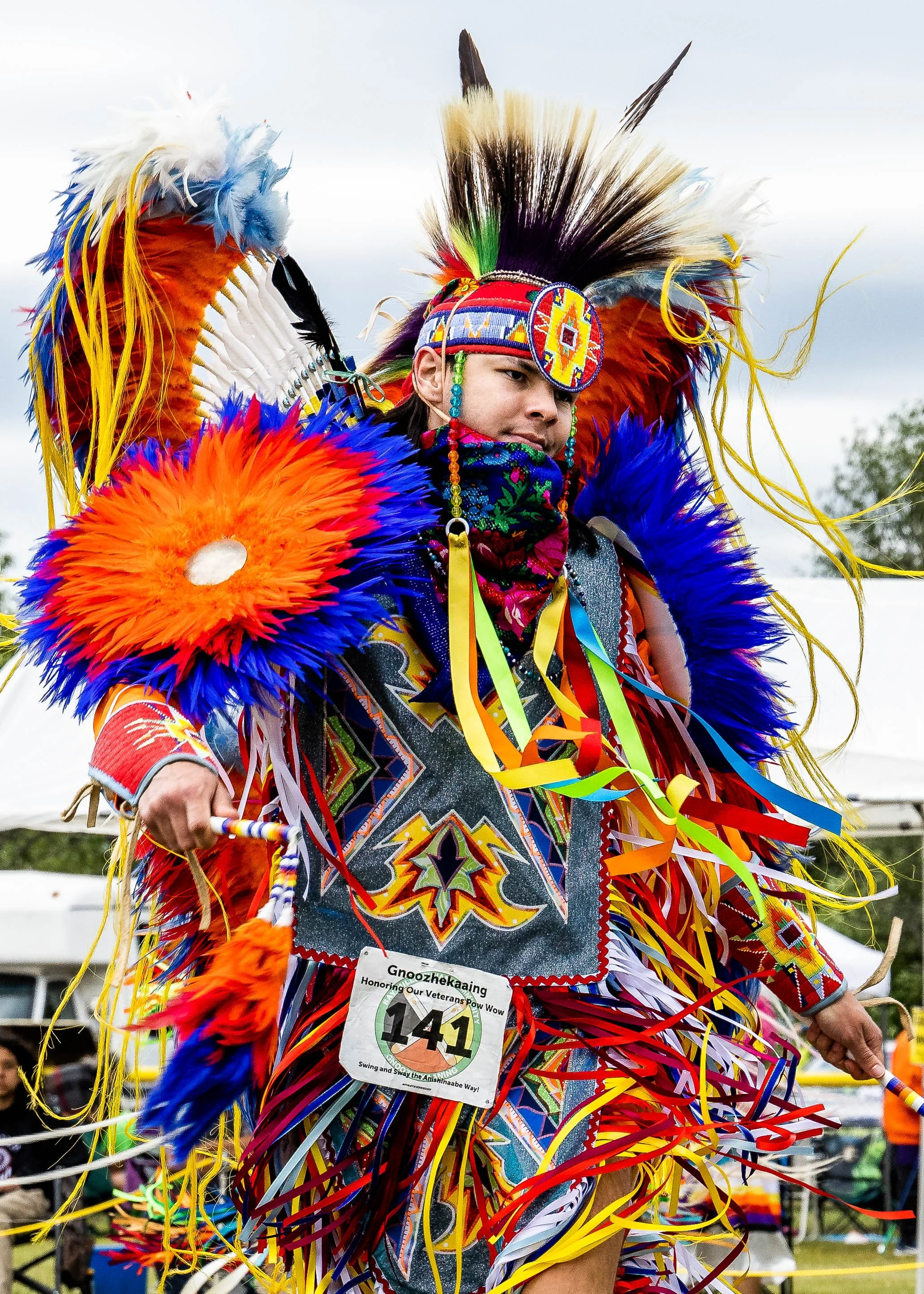 Person dressed in elaborate, colorful traditional Native American regalia with feathers, beadwork, and ribbons, participating in a cultural event or celebration.