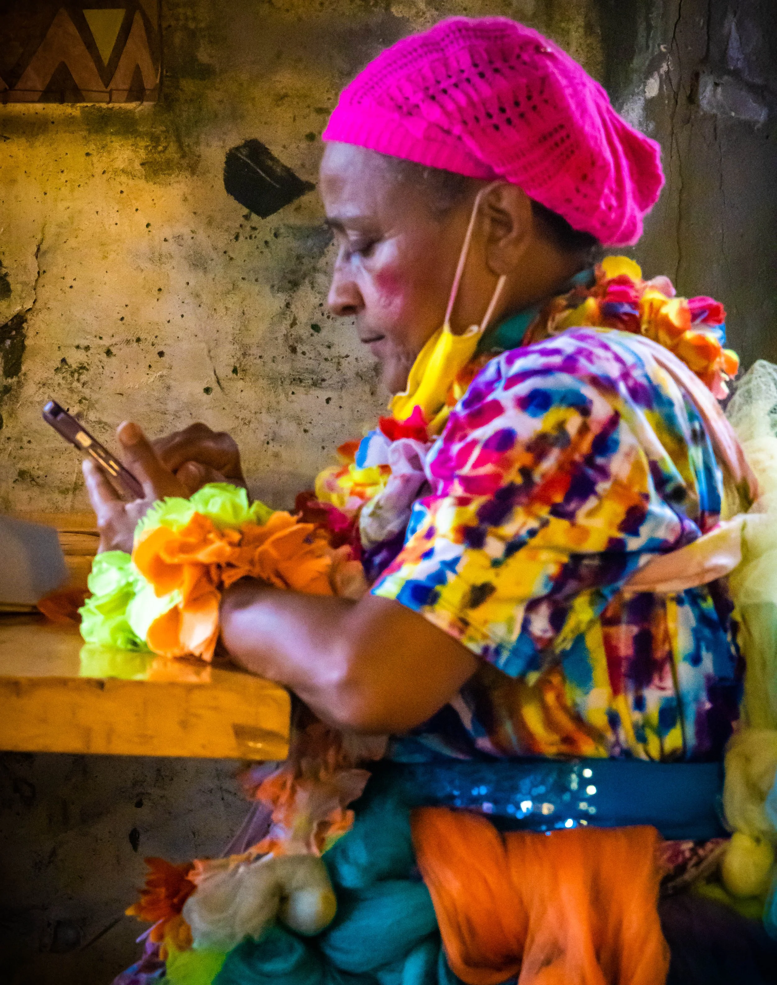 A person dressed in colorful, festive clothing and accessories looking at their phone, wearing a pink knitted hat, surrounded by bright flowers, with a wall in the background.