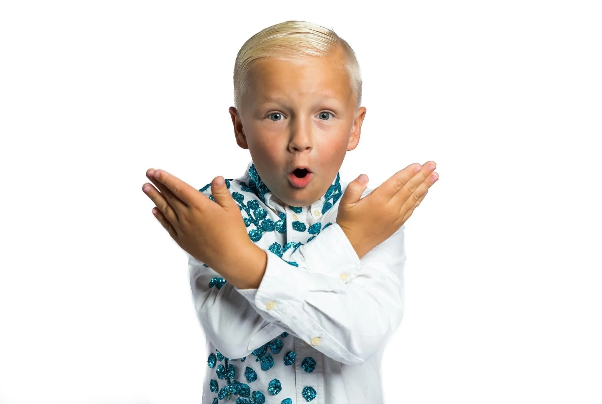 A young boy with platinum blonde hair and blue eyes, wearing a white shirt with turquoise sequined paisley pattern, crossed arms, and surprised expression.