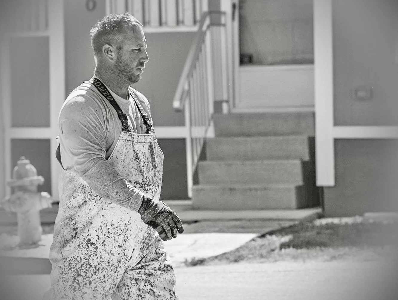 A man with a beard wearing work clothes, including a paint-splattered apron and gloves, standing outside in black and white.