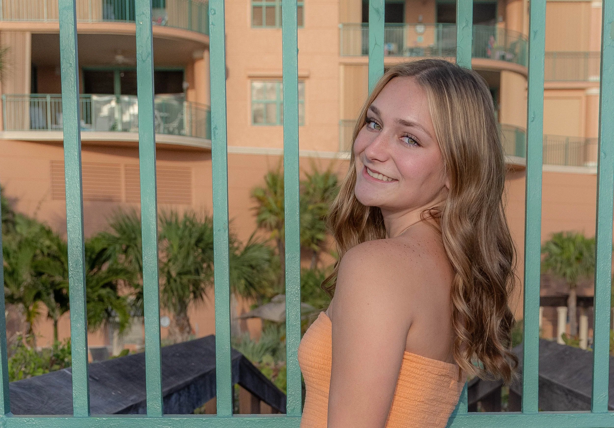 A young woman with long, wavy brown hair and bright blue eyes smiling as she looks over her shoulder, standing behind a turquoise metal fence with an apartment building in the background.
