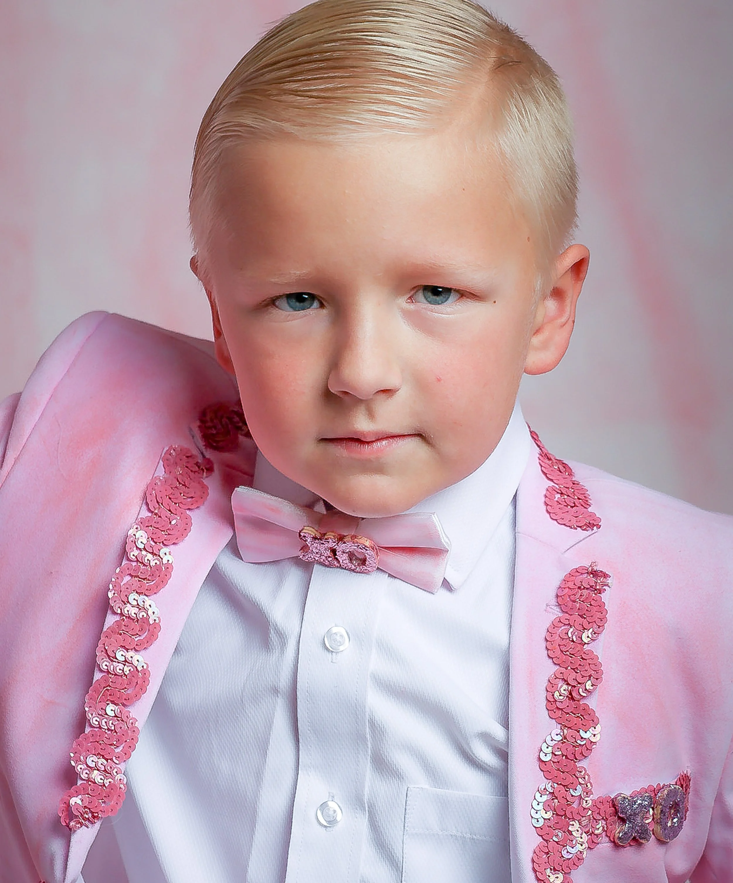 Close-up of a young boy with blonde hair and blue eyes, dressed in formal attire with a pink sequined jacket, white shirt, and pink bow tie, posing against a soft pastel background.
