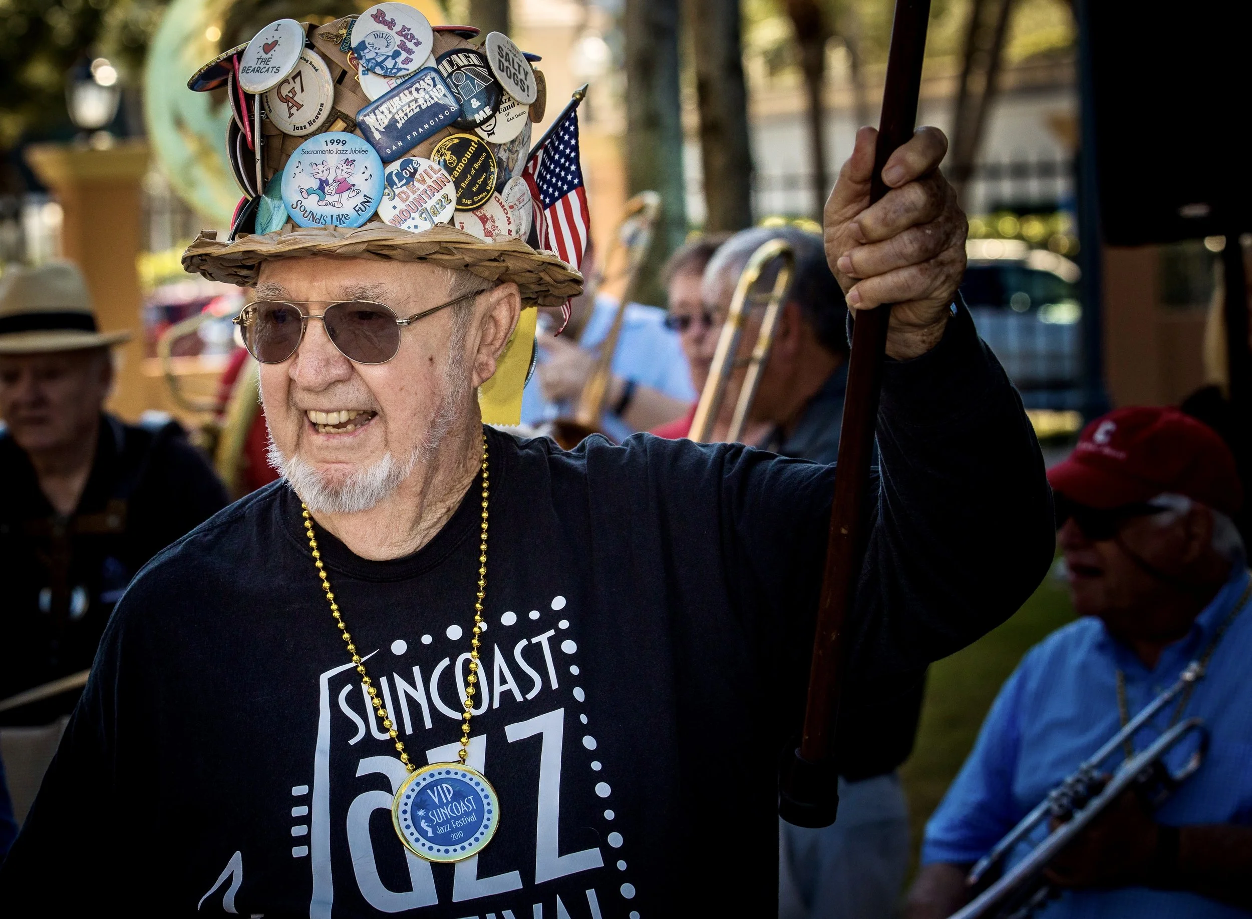 A smiling older man with sunglasses and a white beard wearing a novelty hat decorated with various patches and stickers, including one that says 'The Bearcats' and others referencing jazz and a 1999 festival. He is wearing a black T-shirt with white 