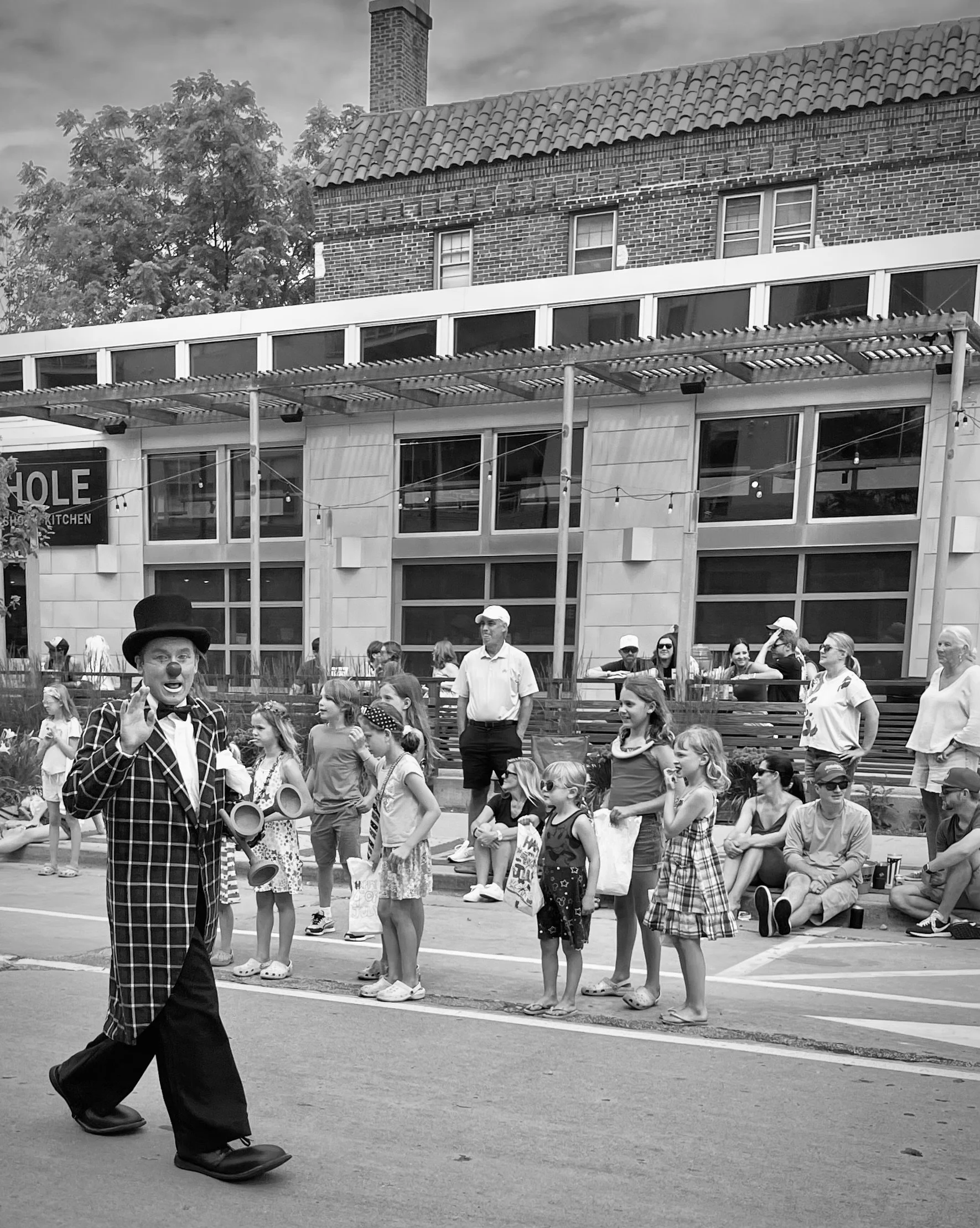 A clown dressed in a black and white checkered suit with a bow tie and bowler hat, performing or entertaining a group of children and adults in an outdoor area, with a building and trees in the background.