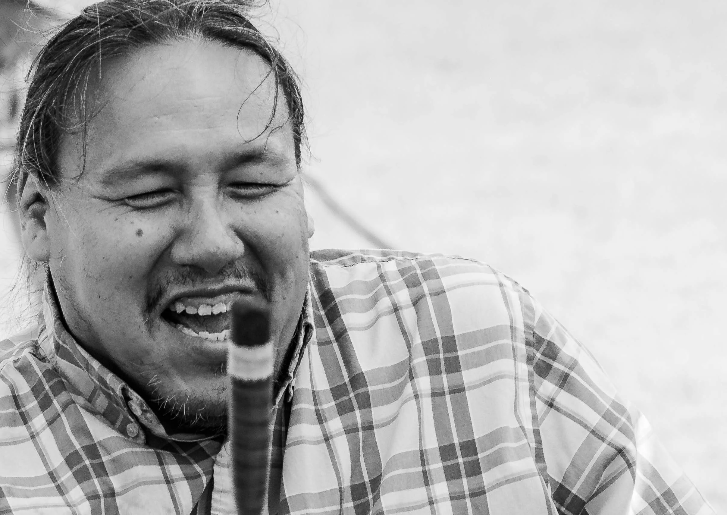 A person smiling broadly, wearing a checkered shirt, with a dark object or marker in the foreground, and a plain light background.