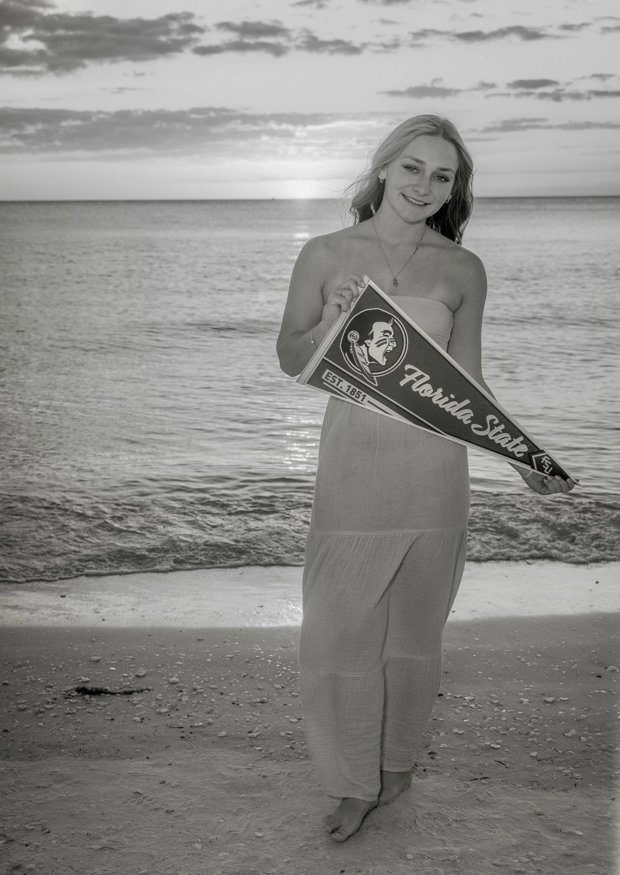 A young woman in a strapless dress holding a Florida State pennant on a beach at sunset, with the ocean and partly cloudy sky in the background.