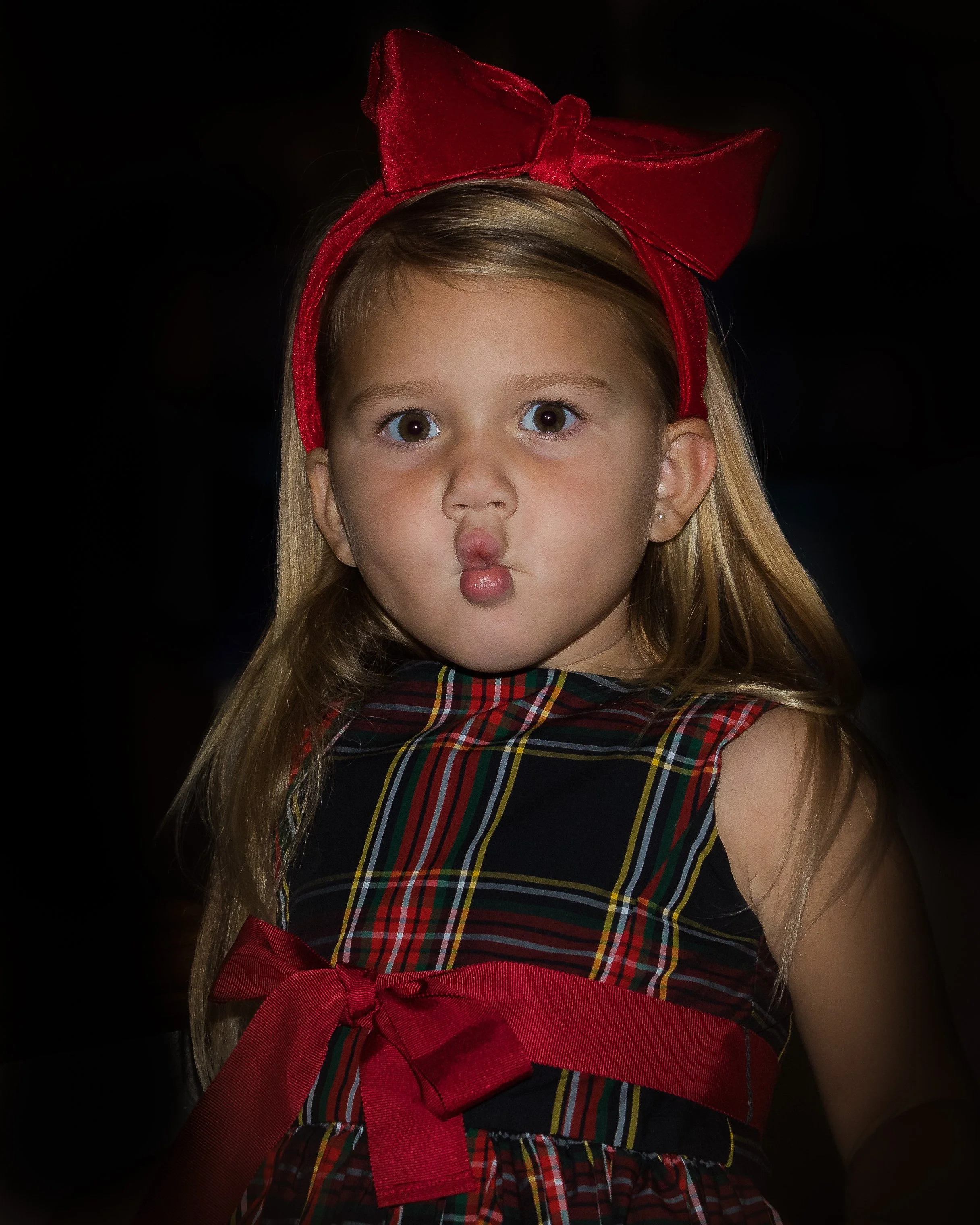 Close-up of a young girl with long blonde hair and wide eyes, wearing a red bow headband and a dress with a tartan pattern, against a black background.