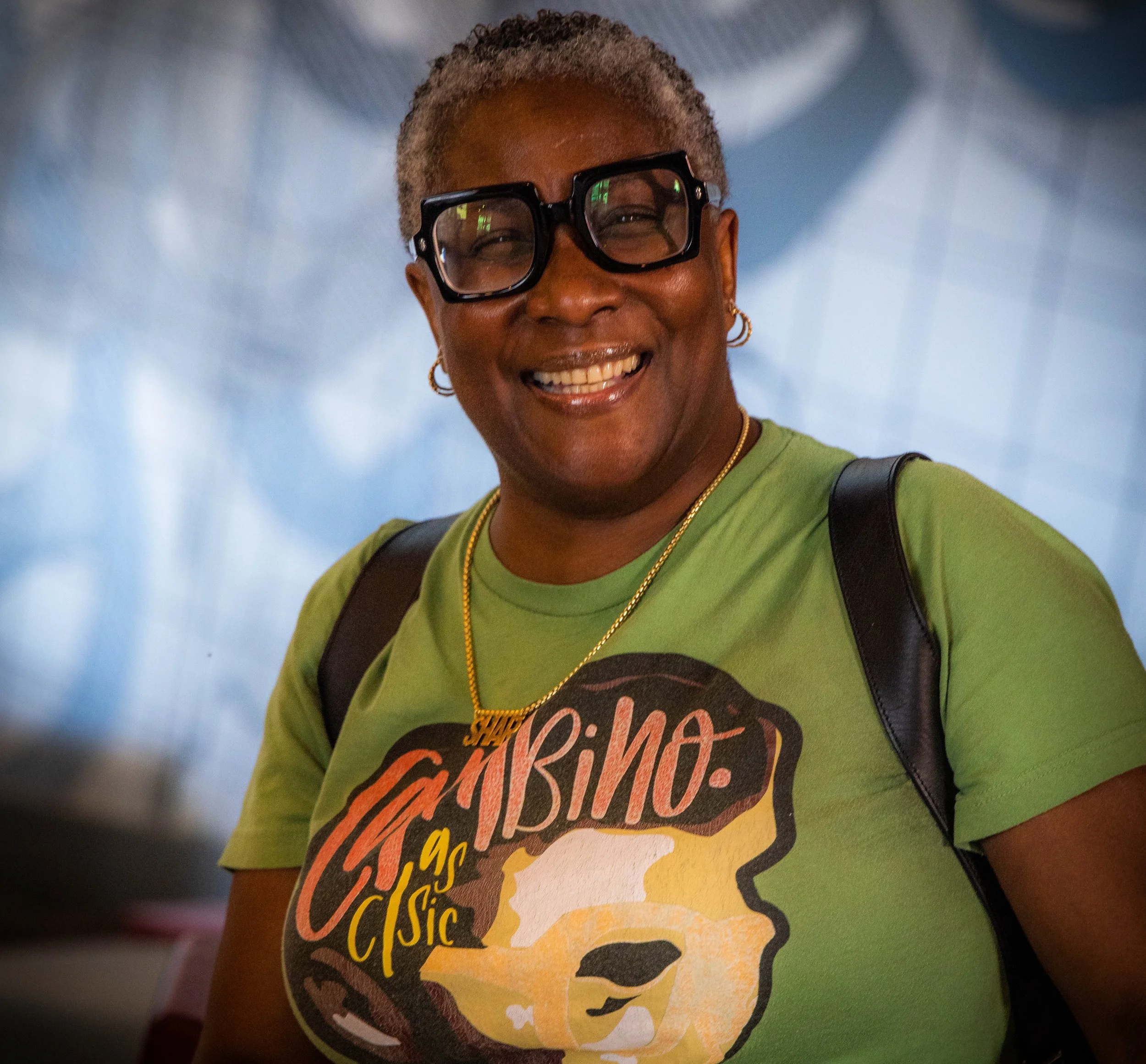 Close-up of a smiling woman with short gray hair, wearing large black glasses, a green shirt, gold jewelry, and carrying a black strap, with a blurred background.