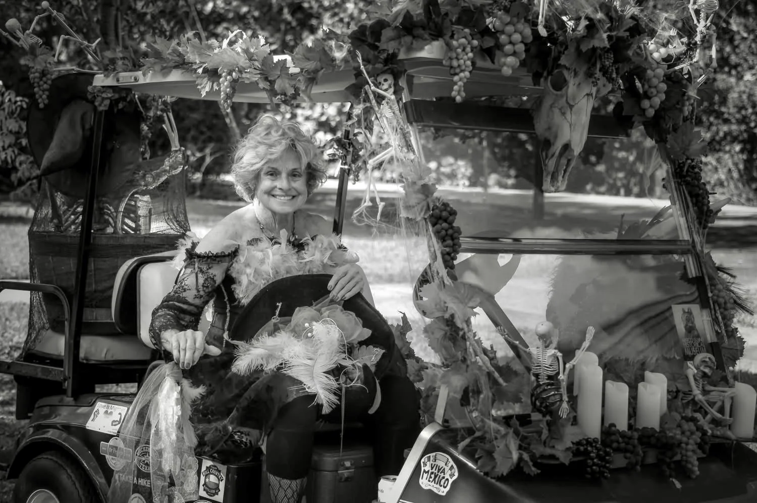 An elderly woman dressed in festive, old-fashioned attire sitting in a decorated cart. The cart is adorned with grapes, candles, and skeleton decorations, suggesting a celebration or parade, possibly Halloween or Día de los Muertos.
