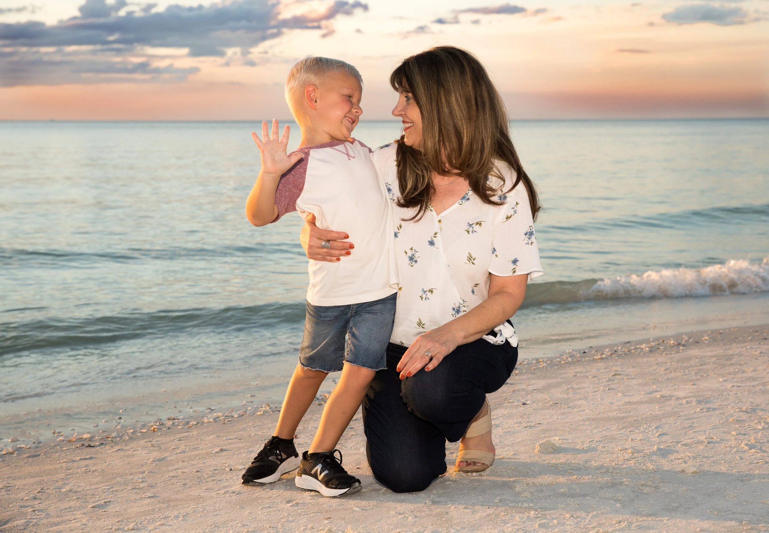 A woman kneeling on the beach holding a young boy with blonde hair and a white shirt. They are smiling at each other with the ocean and sunset in the background.