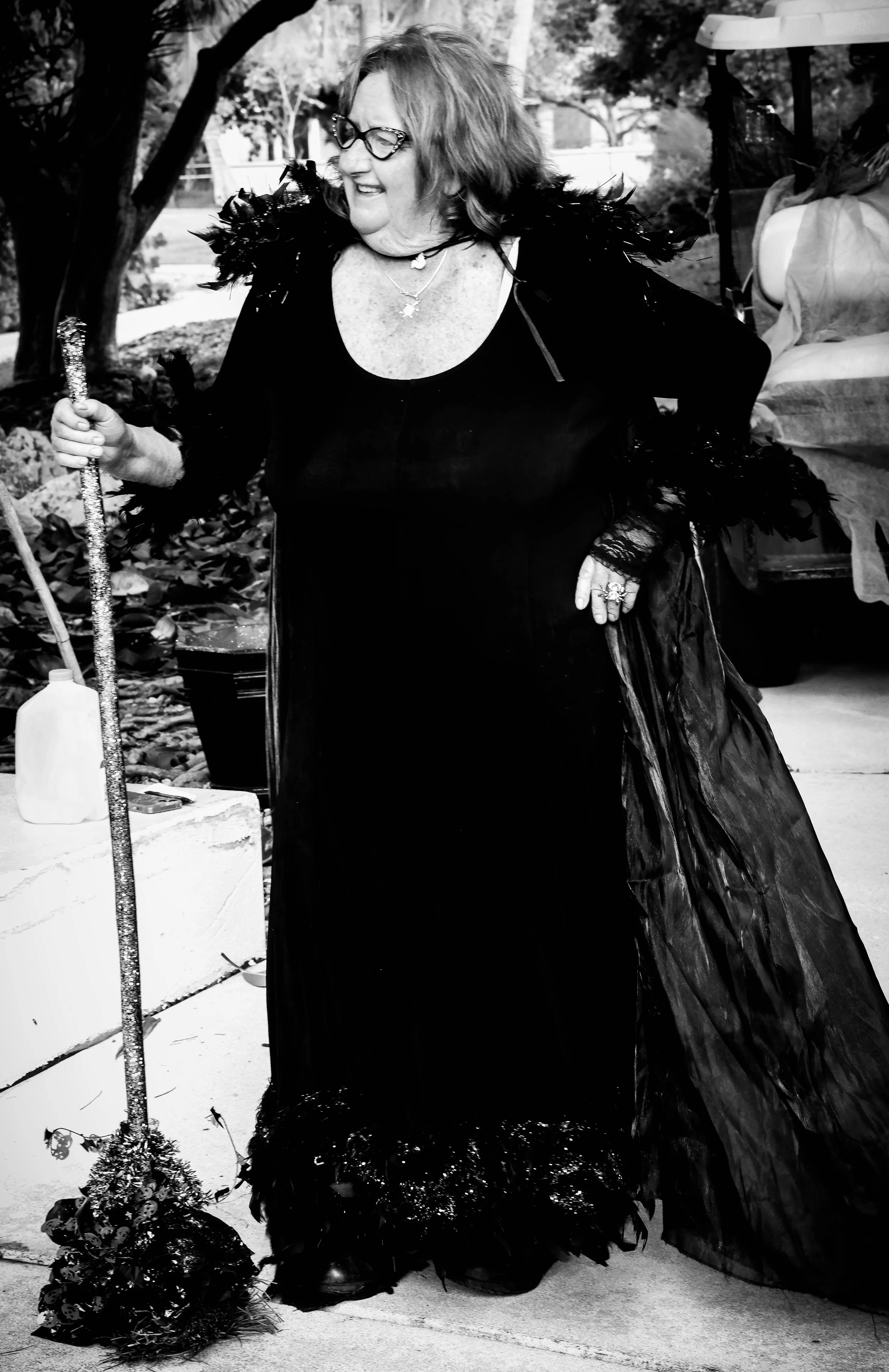 A woman with glasses and musical.ly earrings in a long dark gown with feathered details, holding a sparkly staff, standing outdoors with trees in the background. She is smiling and wearing layered necklaces.