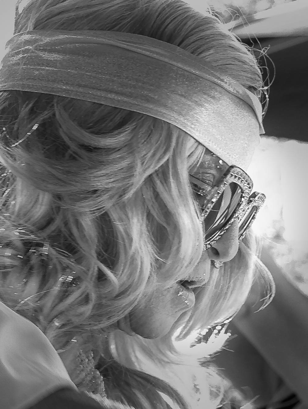 Close-up side profile of a woman with long, curly hair, wearing sunglasses, a headband, and a shiny scarf, looking to the right.