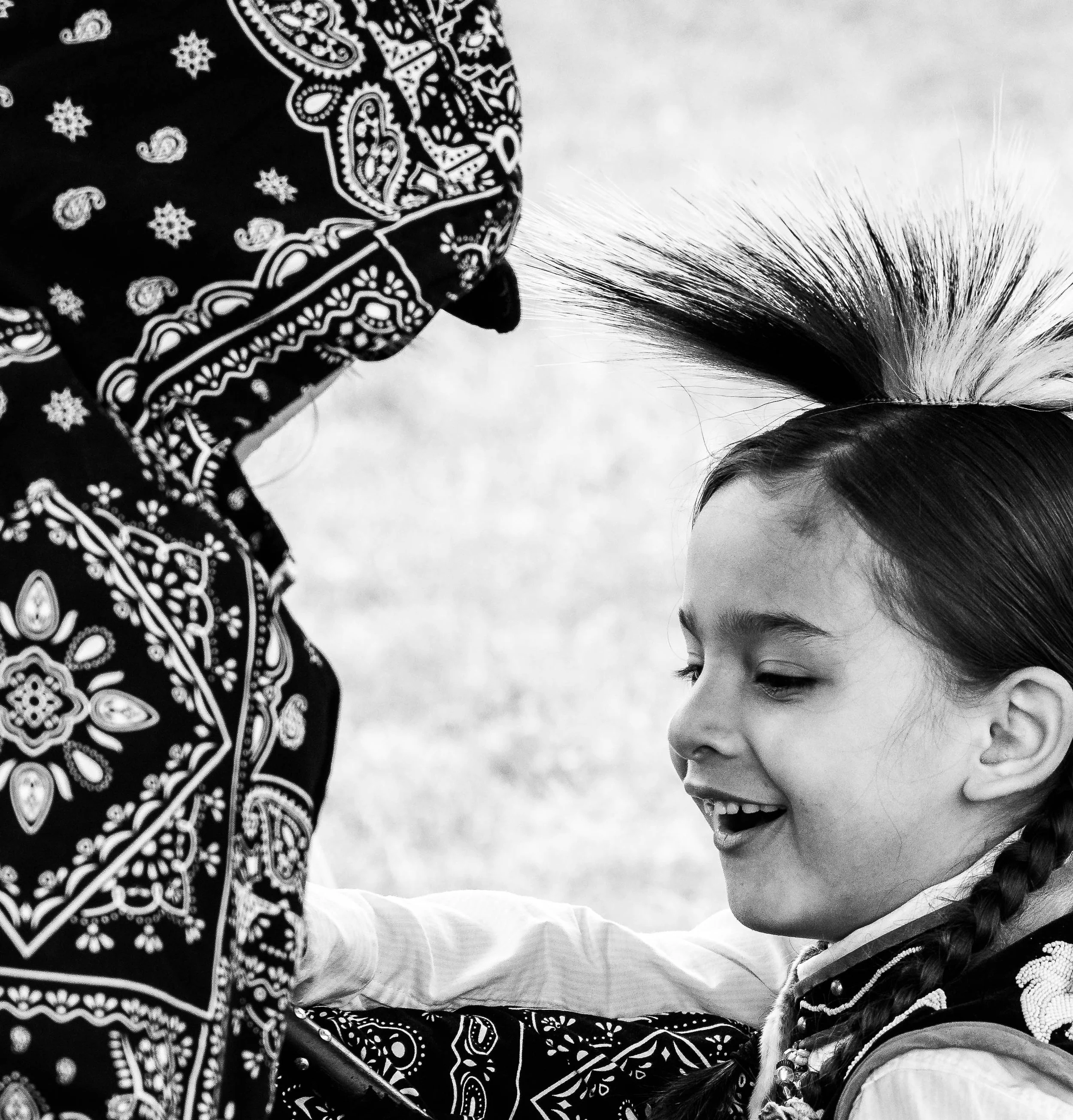 A young girl with her eyes closed smiling, wrapped in a patterned blanket or shawl, with an adult's arm nearby, both outdoors.