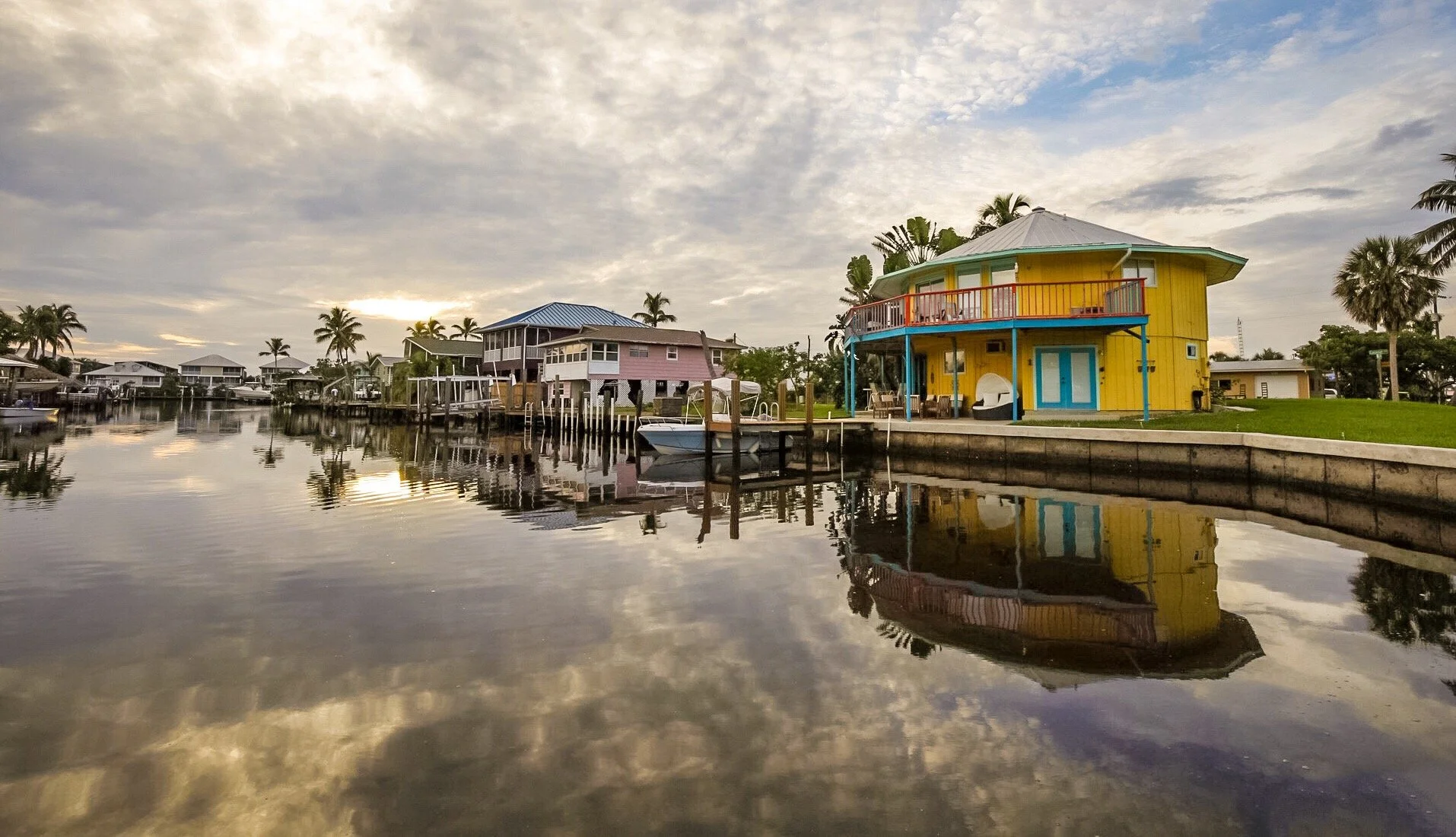 A colorful house with yellow walls, blue door, and red and blue balcony, situated by a calm water canal with reflections of the house and sky, in a tropical area with palm trees and other houses in the background, under a cloudy sky.