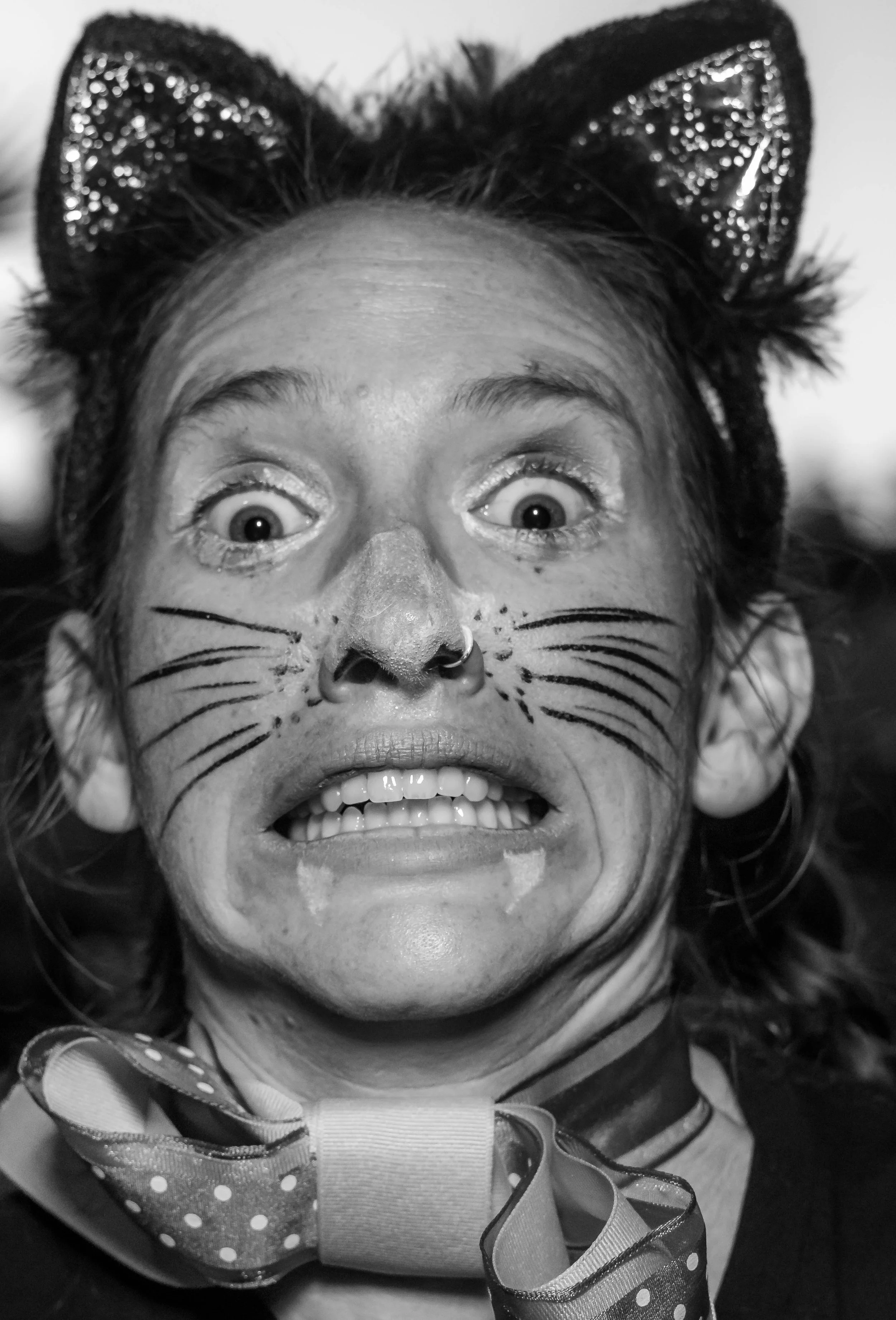 A person with a wide smile, face painted with a cat nose and whiskers, wearing cat ears headband with sequins, and a large polka dot bow with ribbon around the neck, in a playful pose.