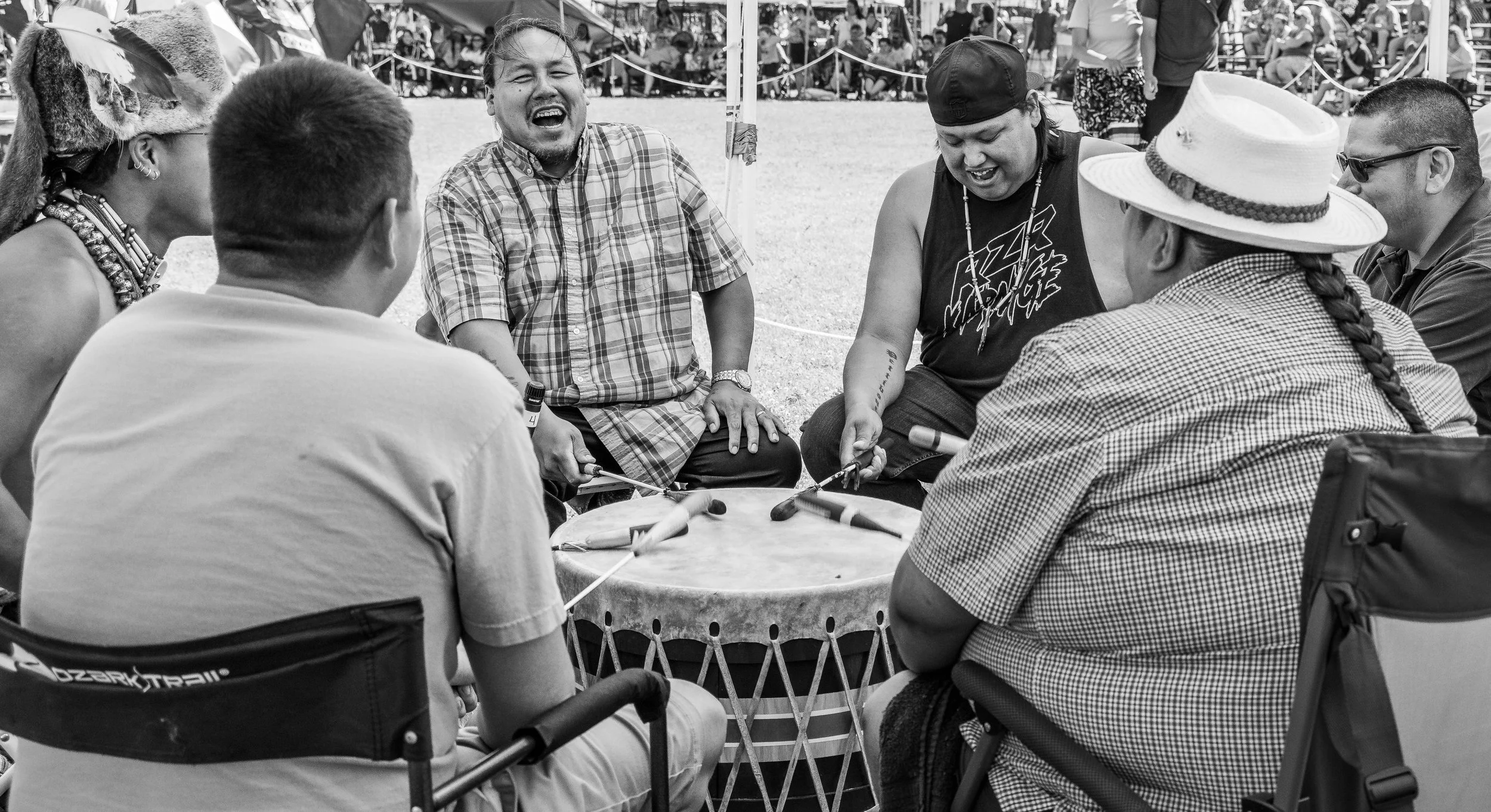 Group of diverse people gathered around a drum at an outdoor event, laughing and enjoying themselves, with a crowd of spectators in the background.