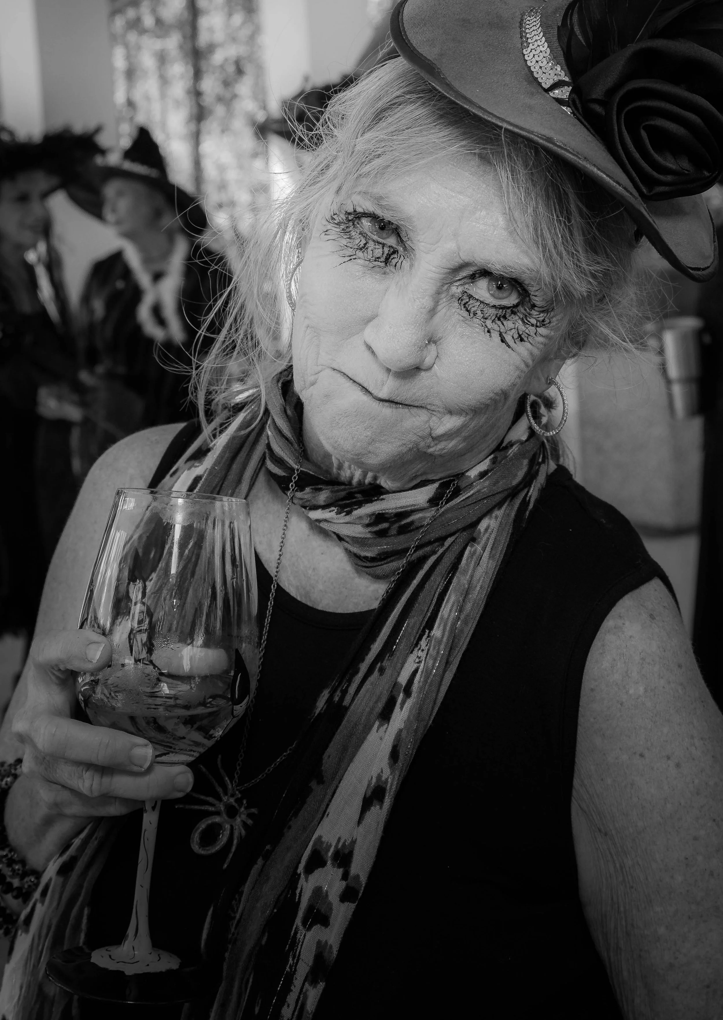 A woman with light-colored hair wearing a hat with a large decorative flower, eye makeup that looks like black tears, and hoop earrings, holding a glass of wine. Two women in similar hats are blurred in the background.