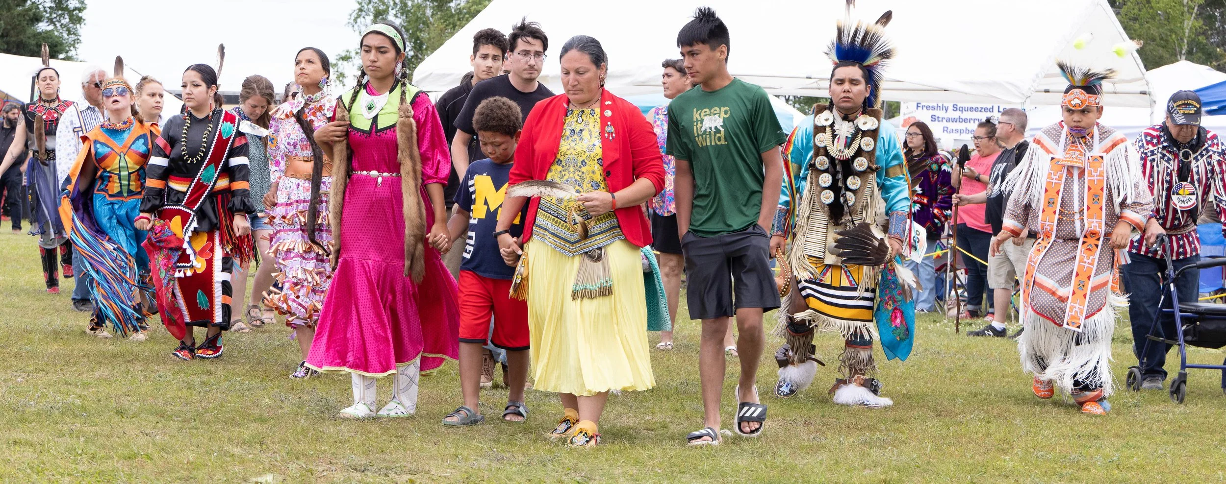 Group of people, including men, women, and children, participating in a cultural event, dressed in traditional Native American clothing, walking together on a grassy field with vendors and tents in the background.